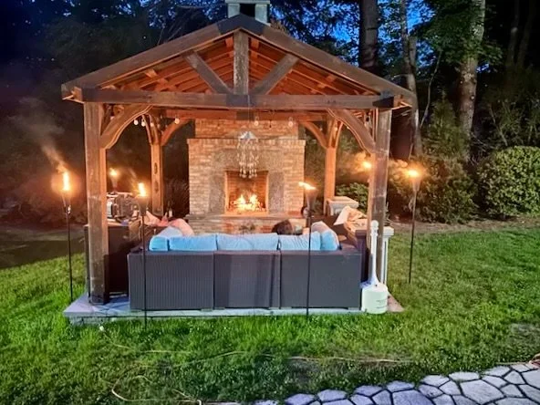 Outdoor gazebo with a stone fireplace, cozy seating, and tiki torches, set in a grassy backyard at dusk for evening gathering