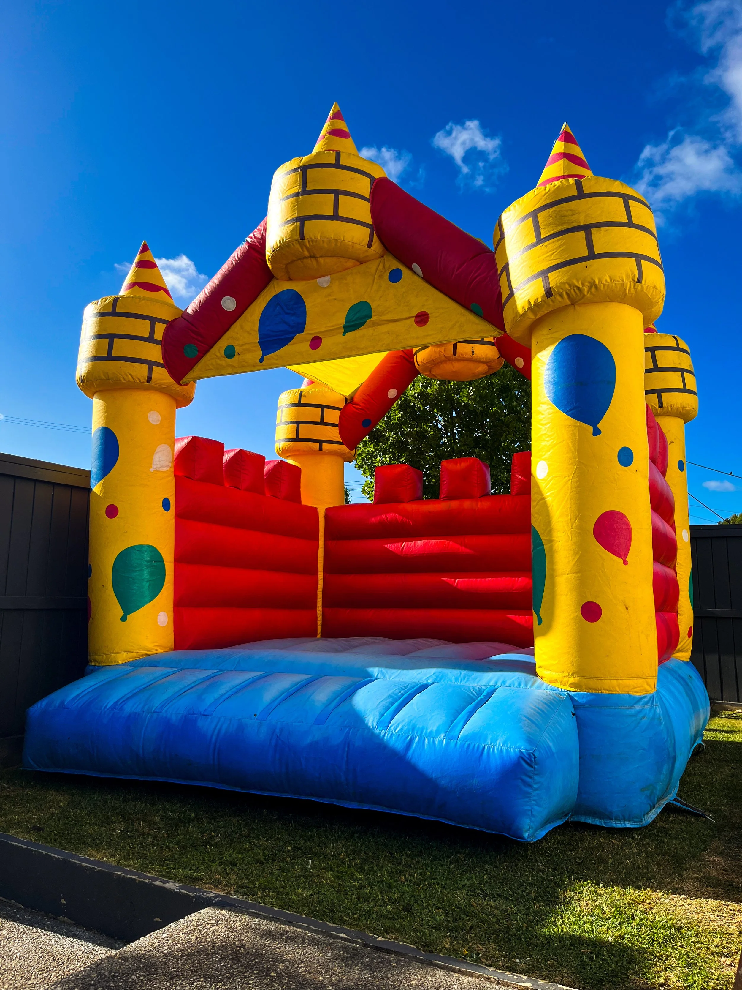 Colorful inflatable bounce house decorated like a castle with yellow, red, and blue colors, topped with cone-shaped turrets and surrounded by a grassy outdoor area under a blue sky.
