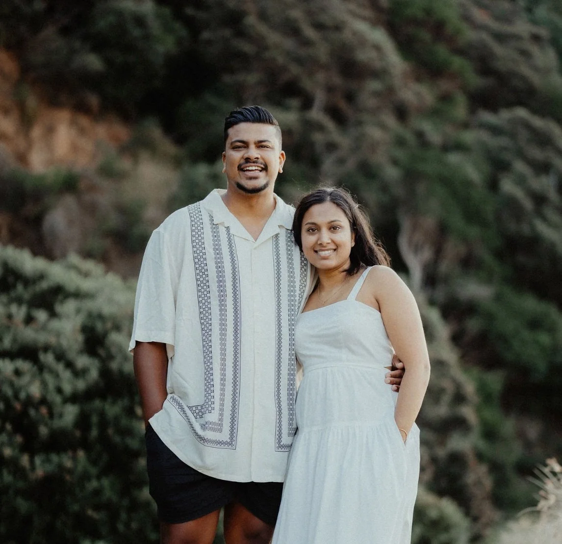 A smiling man and woman in white dresses standing outdoors against a natural background of green bushes and rocky hillside.