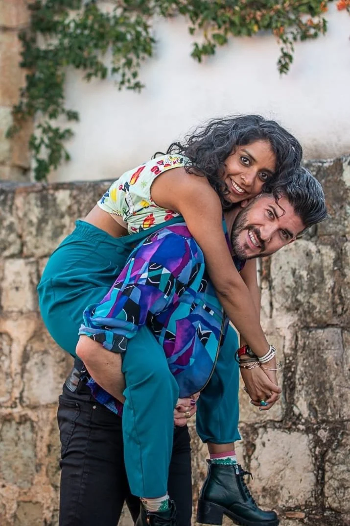 A woman with curly hair wearing a colorful top with cartoon characters and teal pants rides on the back of a man with a beard, wearing a vibrant geometric patterned jacket and teal pants, as they smile and laugh outdoors.