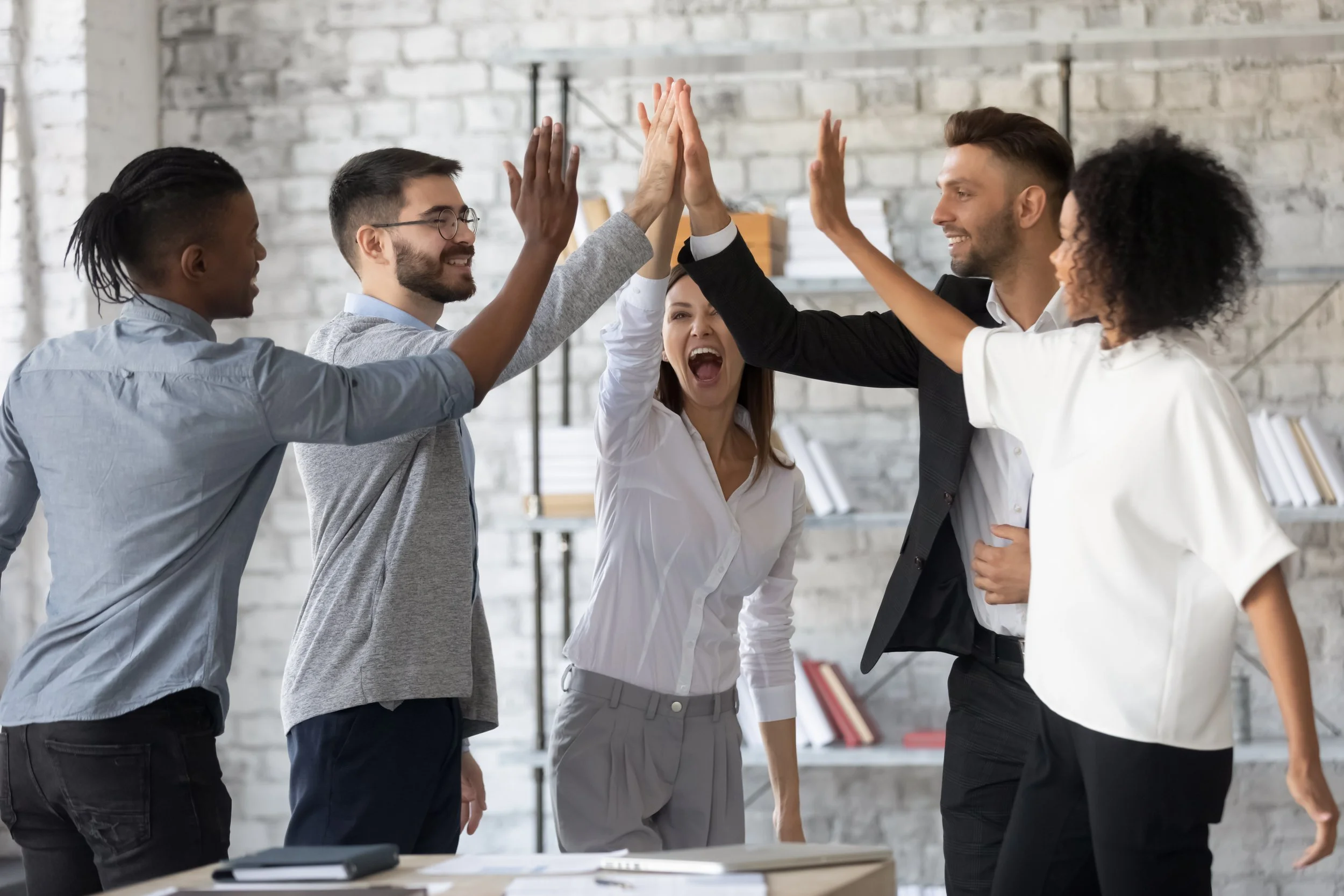 Diverse group of five people in professional attire giving a group high-five in an office setting, showcasing teamwork and success.