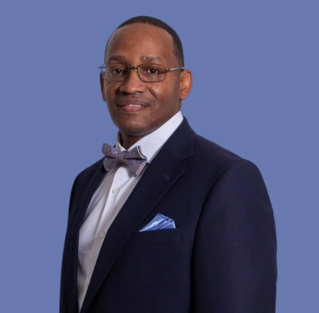 Man in a suit with a bow tie and glasses standing against a blue background.