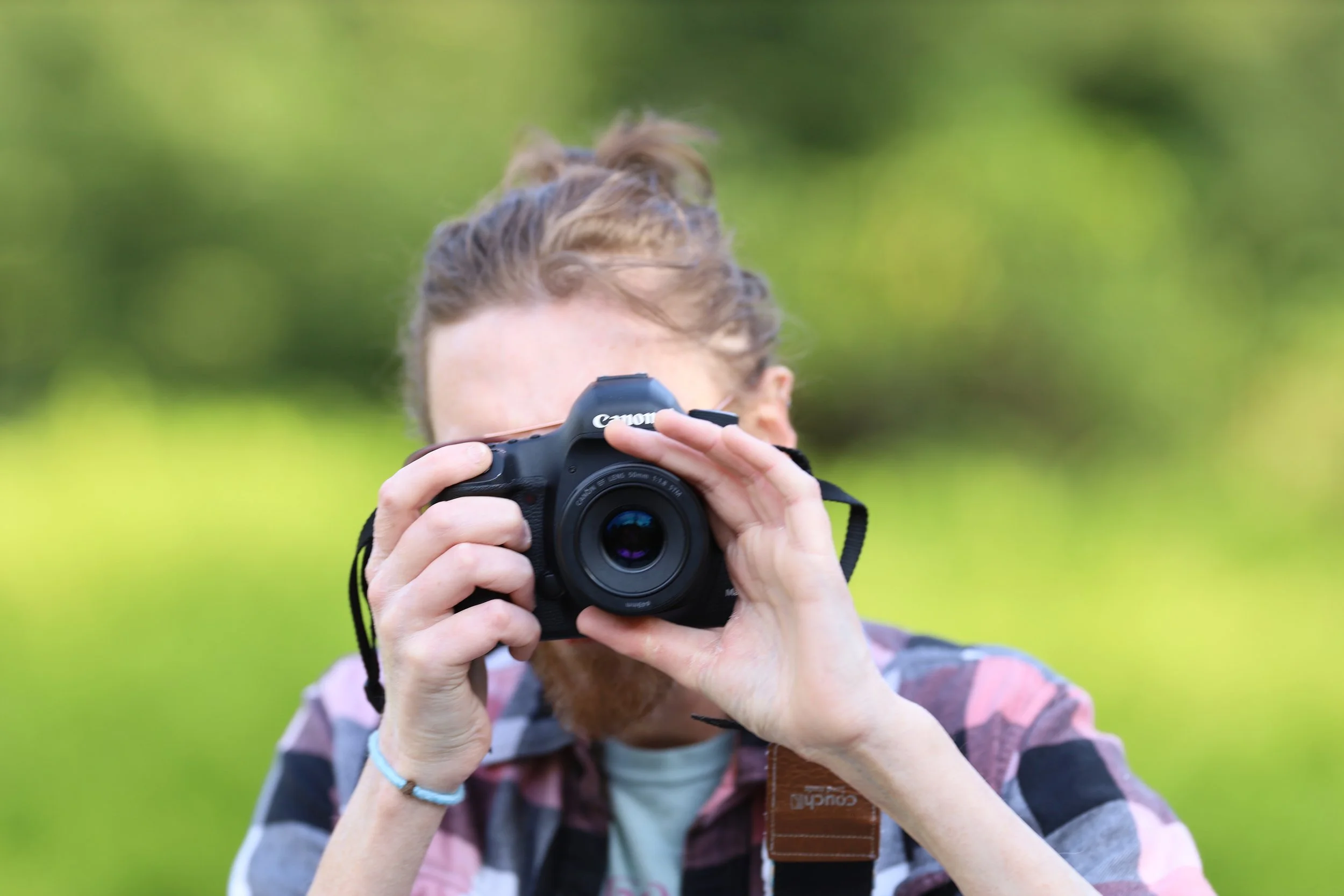 Person holding a Canon camera taking a photo outdoors, wearing a plaid shirt and a bracelet, with a blurred green background.