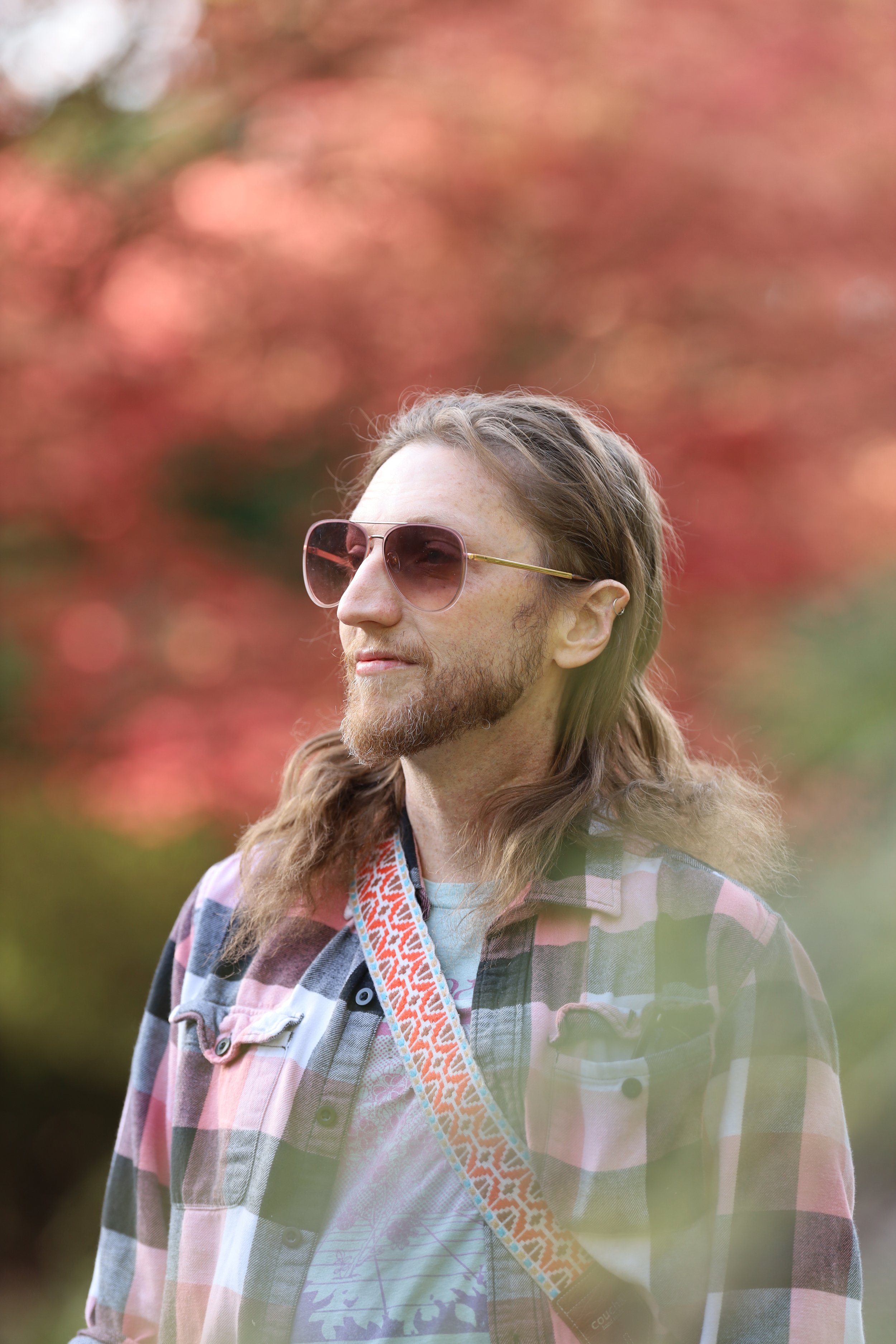 Man with long hair and sunglasses wearing a plaid shirt and patterned strap, standing outdoors with red foliage background.