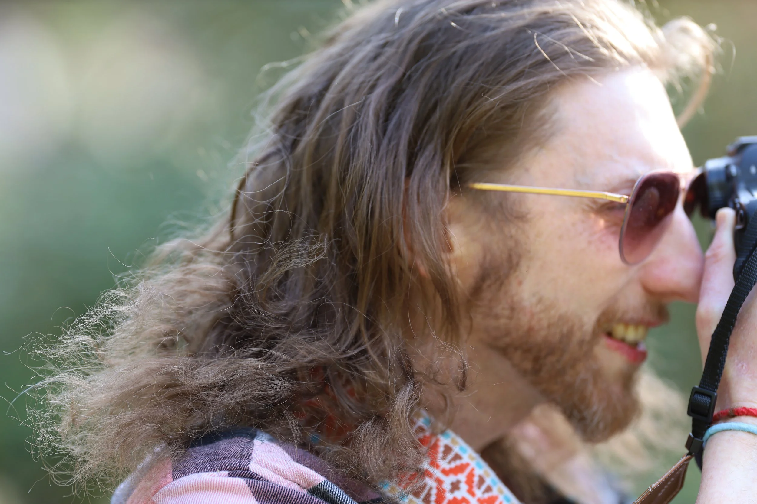 Man with long hair and sunglasses using a camera outdoors.