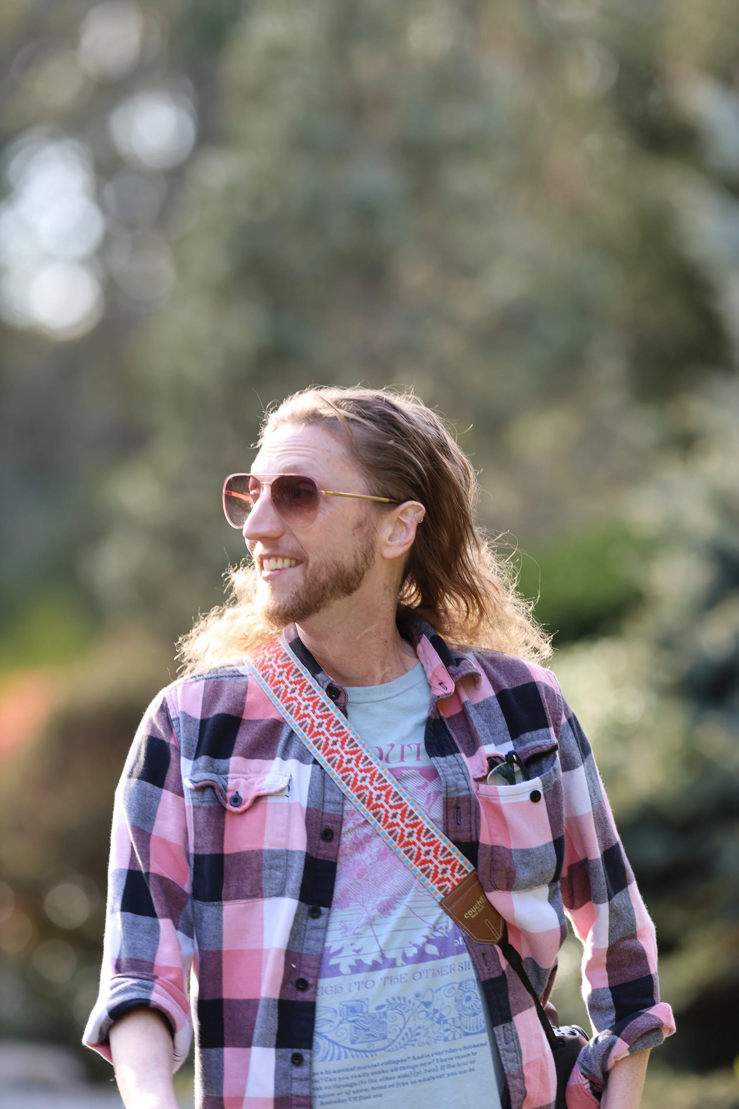 Person wearing a pink plaid shirt and sunglasses, with long hair, smiling outdoors.