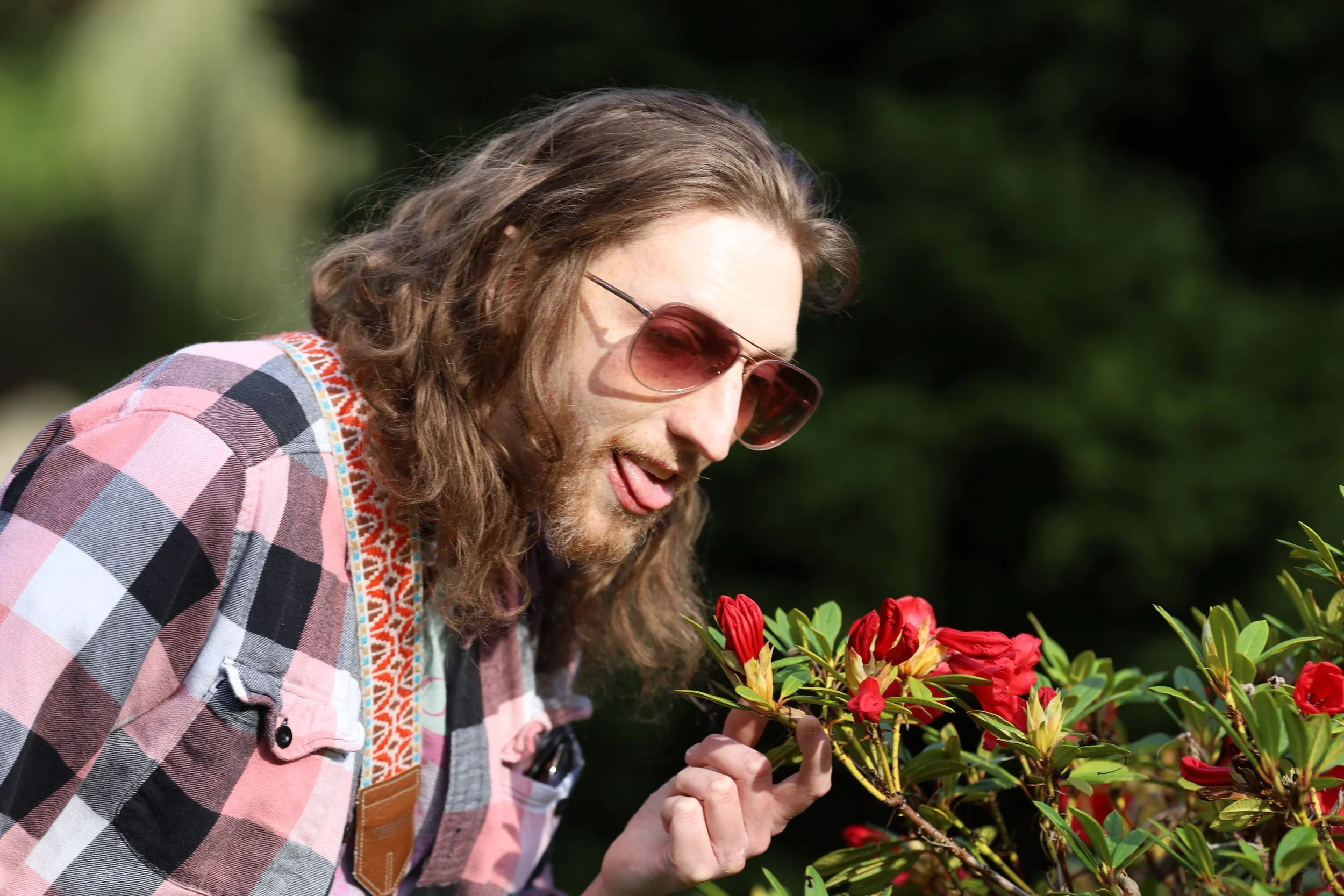 Person with long hair and sunglasses smelling red flowers outdoors