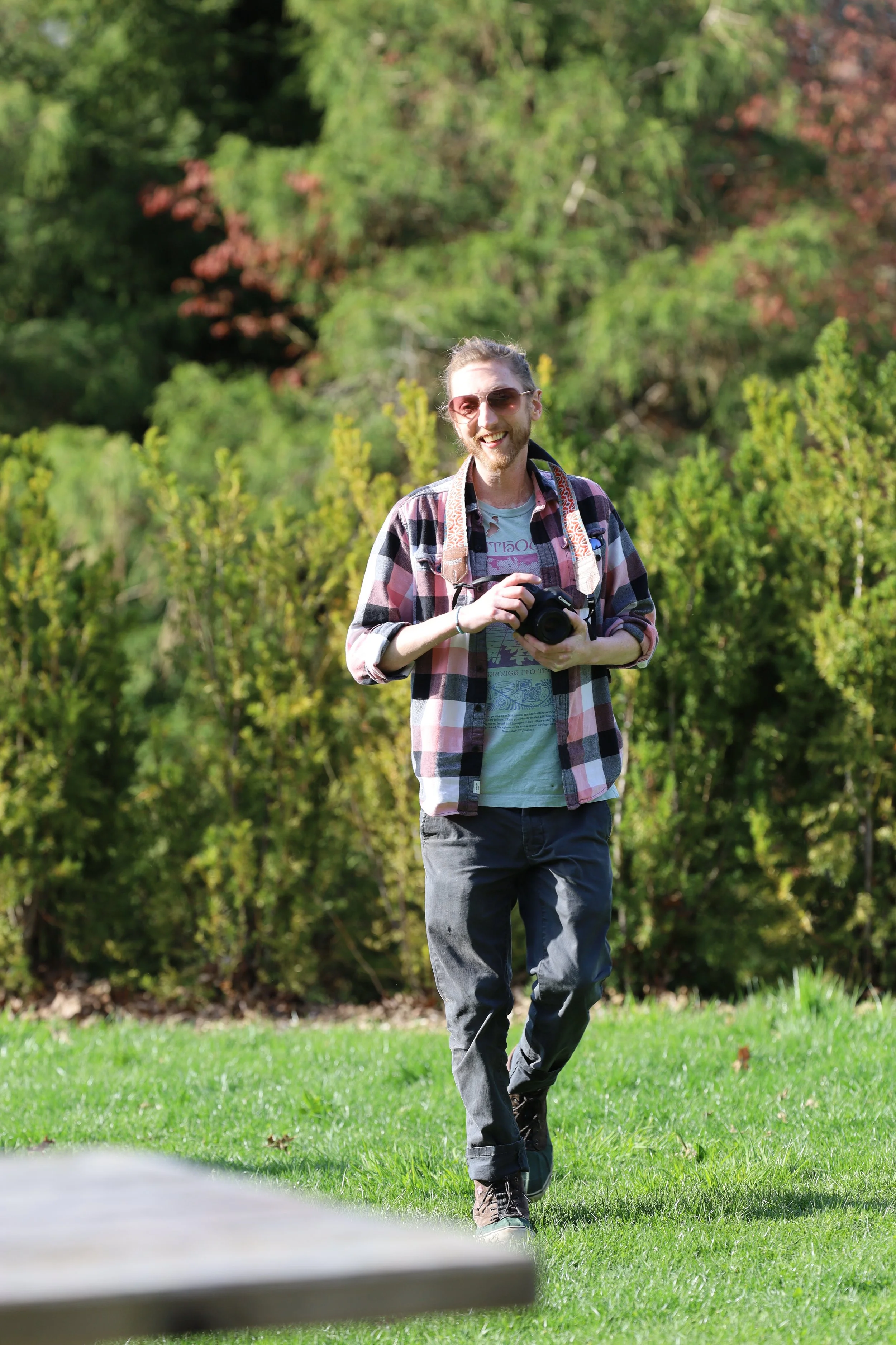 Person in a plaid shirt holding a camera, walking on grass in a park.