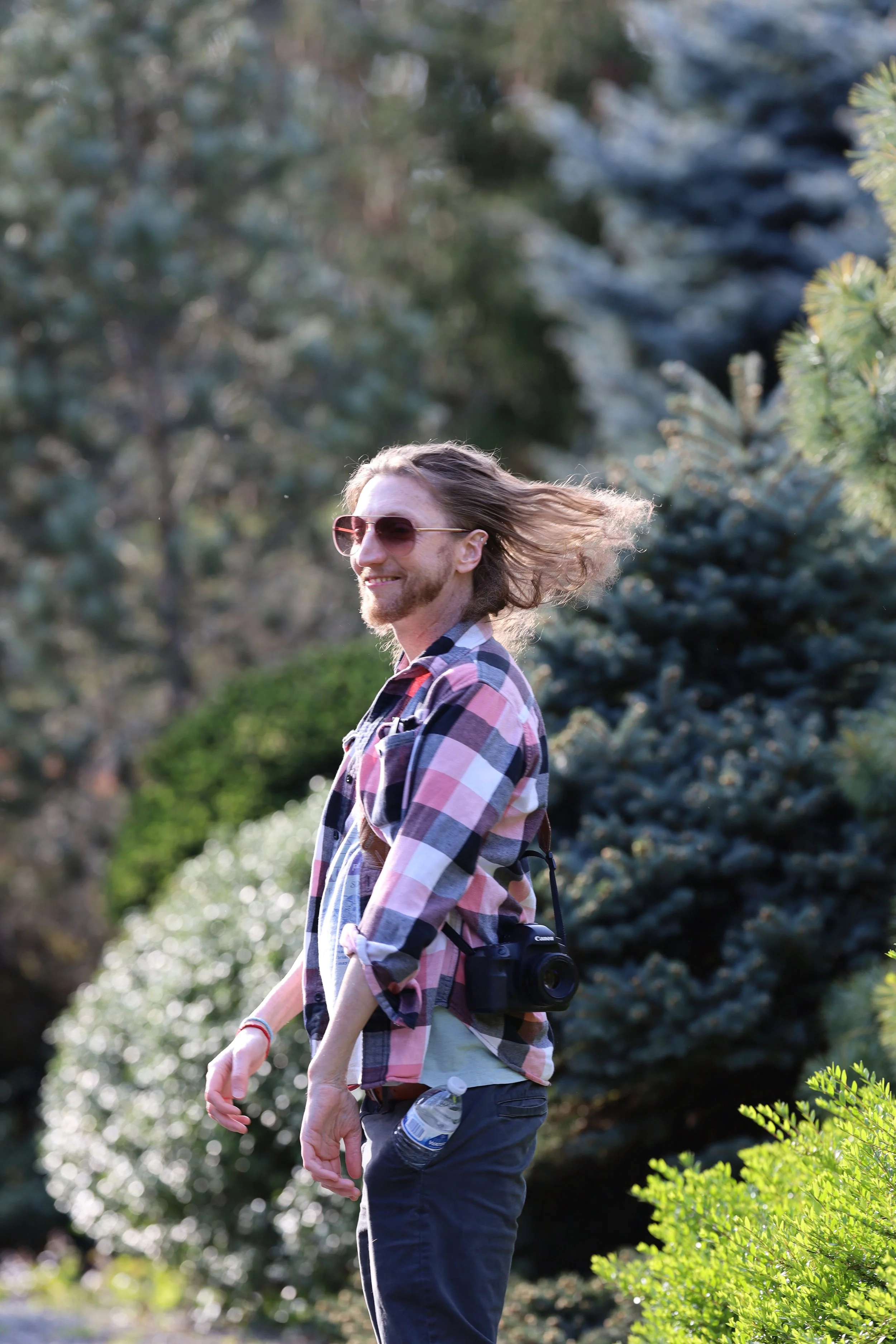 A person with long hair and sunglasses walking outdoors with greenery in the background, wearing a plaid shirt and carrying a camera.