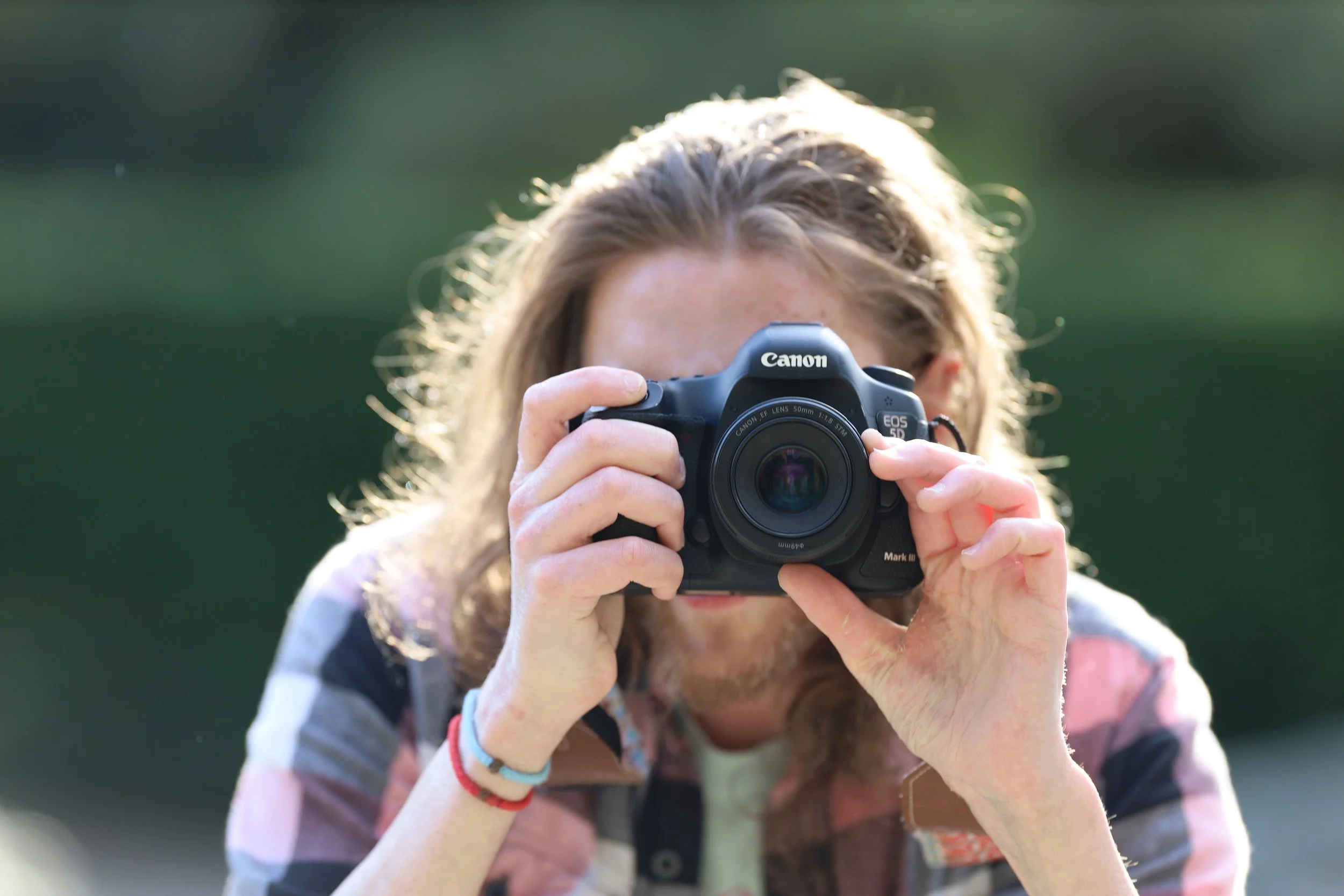 Person holding a Canon EOS 5D Mark III camera outdoors, pointing towards the viewer.