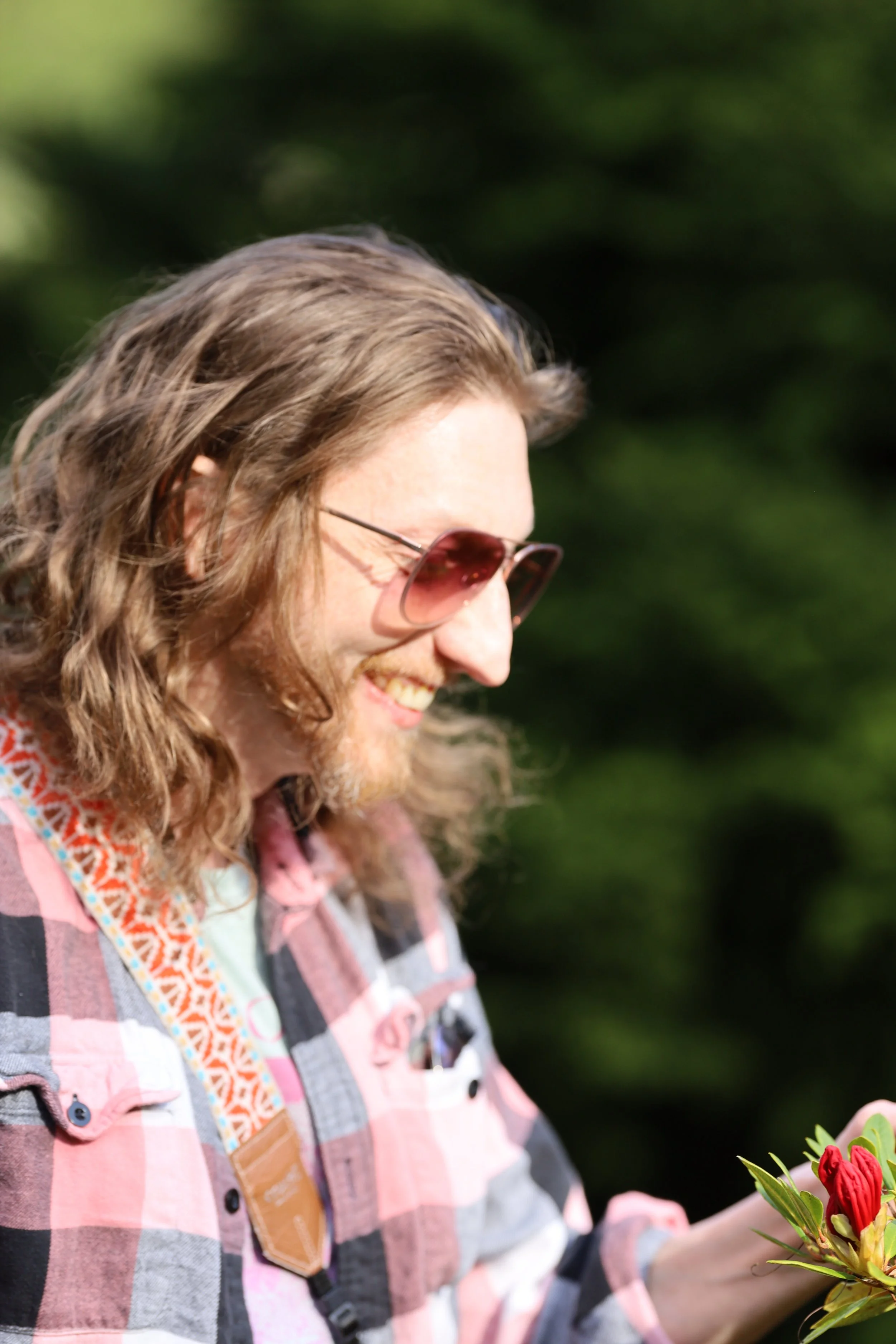 Man with long hair and sunglasses smiling, wearing a plaid shirt, holding flowers.