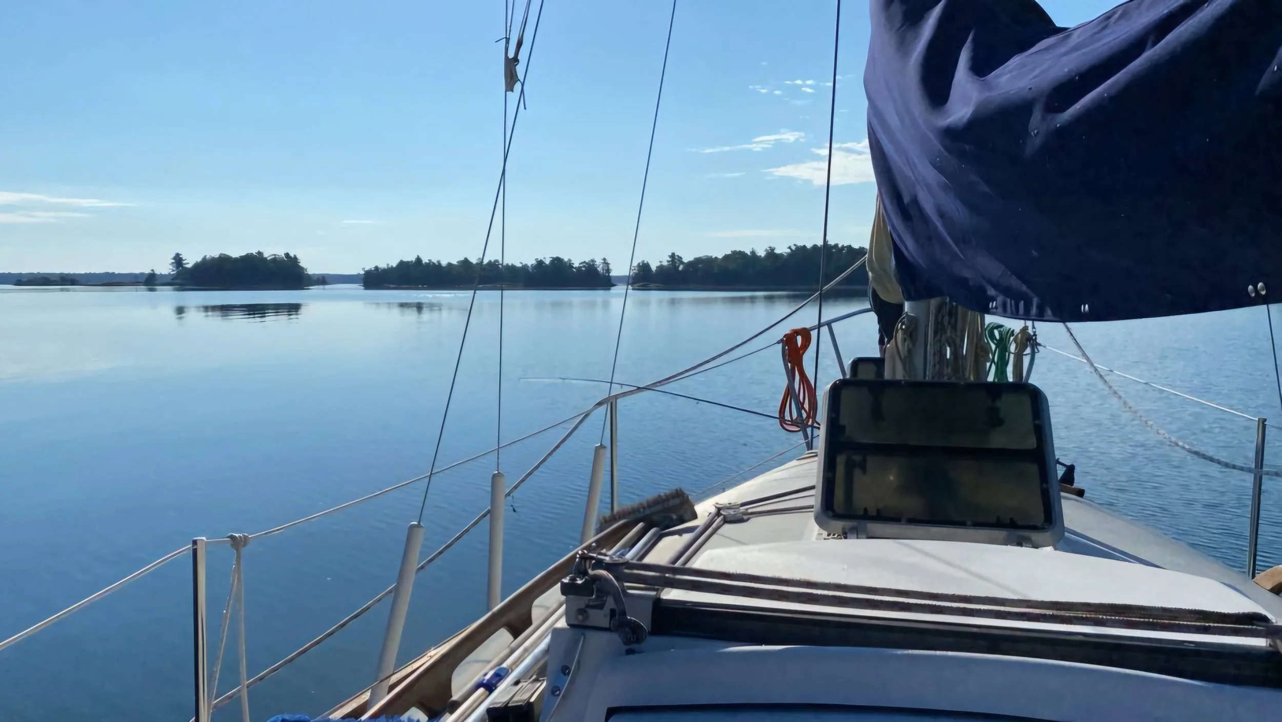 View from a sailboat on a calm lake with distant islands under a clear blue sky.
