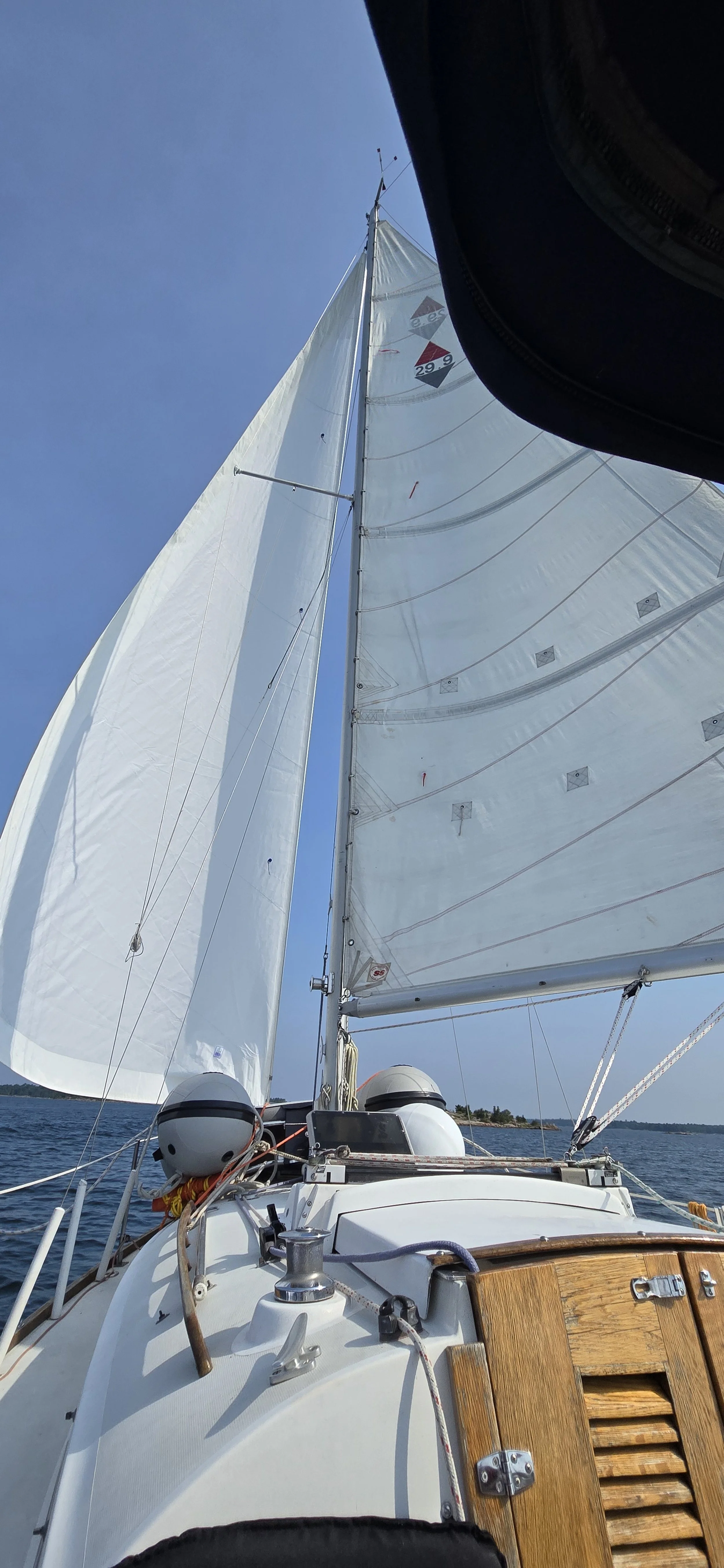 View from the deck of a sailboat looking up at the white sails against a clear blue sky. The boat's deck features ropes and wooden elements.