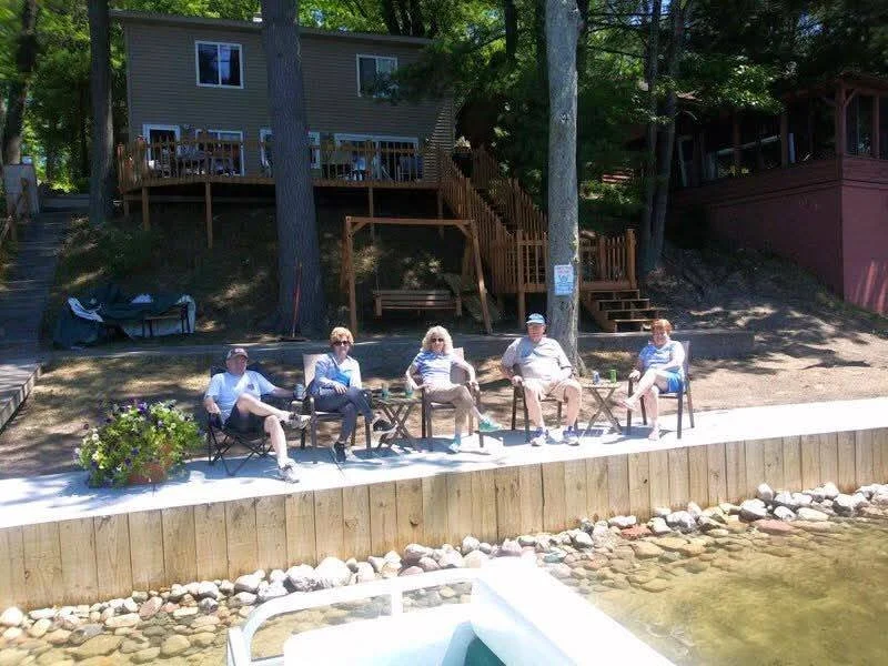 Five people sitting on chairs along a lakeside deck, with stacked rocks and water in the foreground and trees and houses in the background.