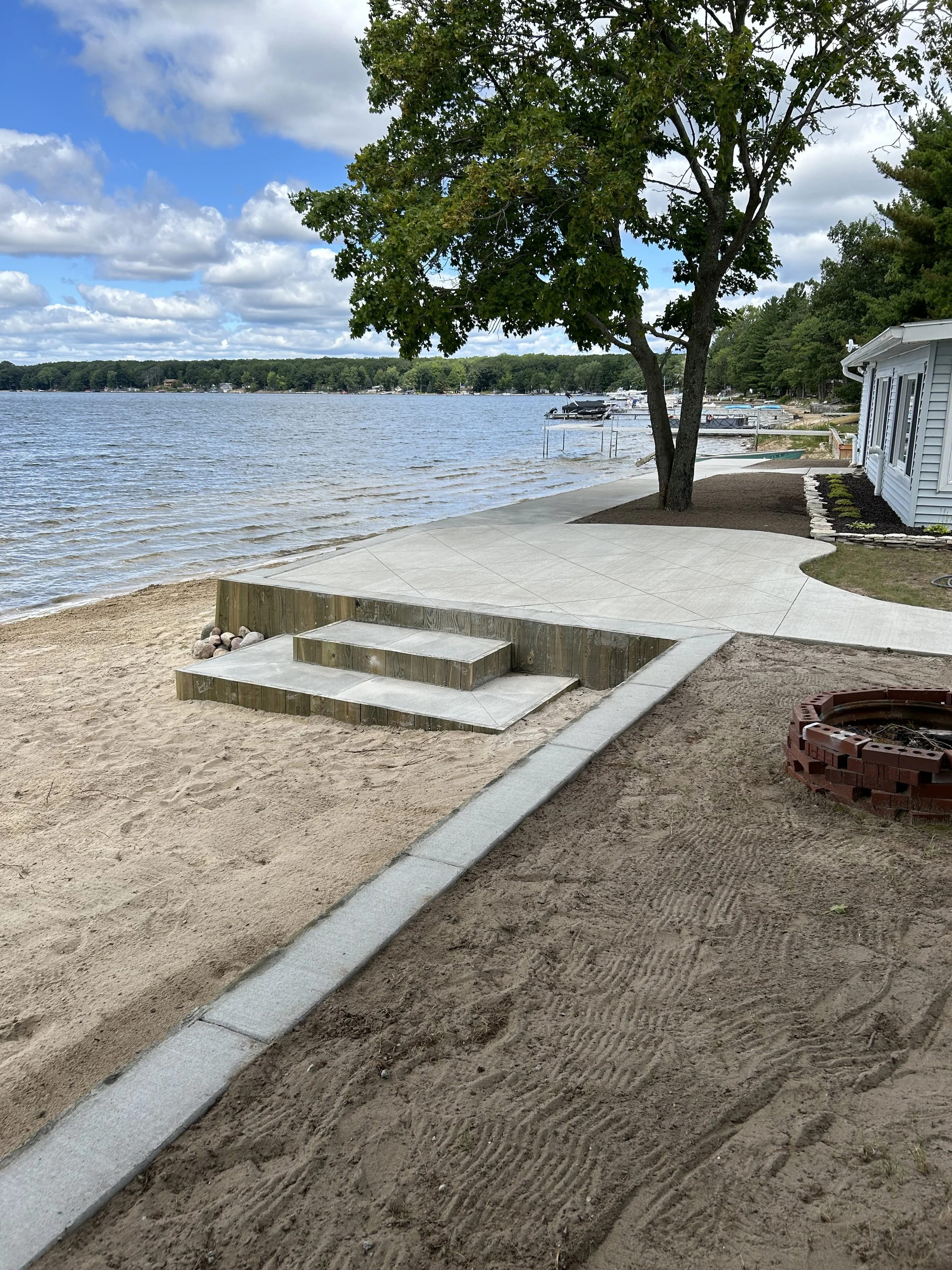 A lakeside yard with a large tree, boats docked along the water, a house with white siding, a concrete walkway, and a fire ring in the dirt.