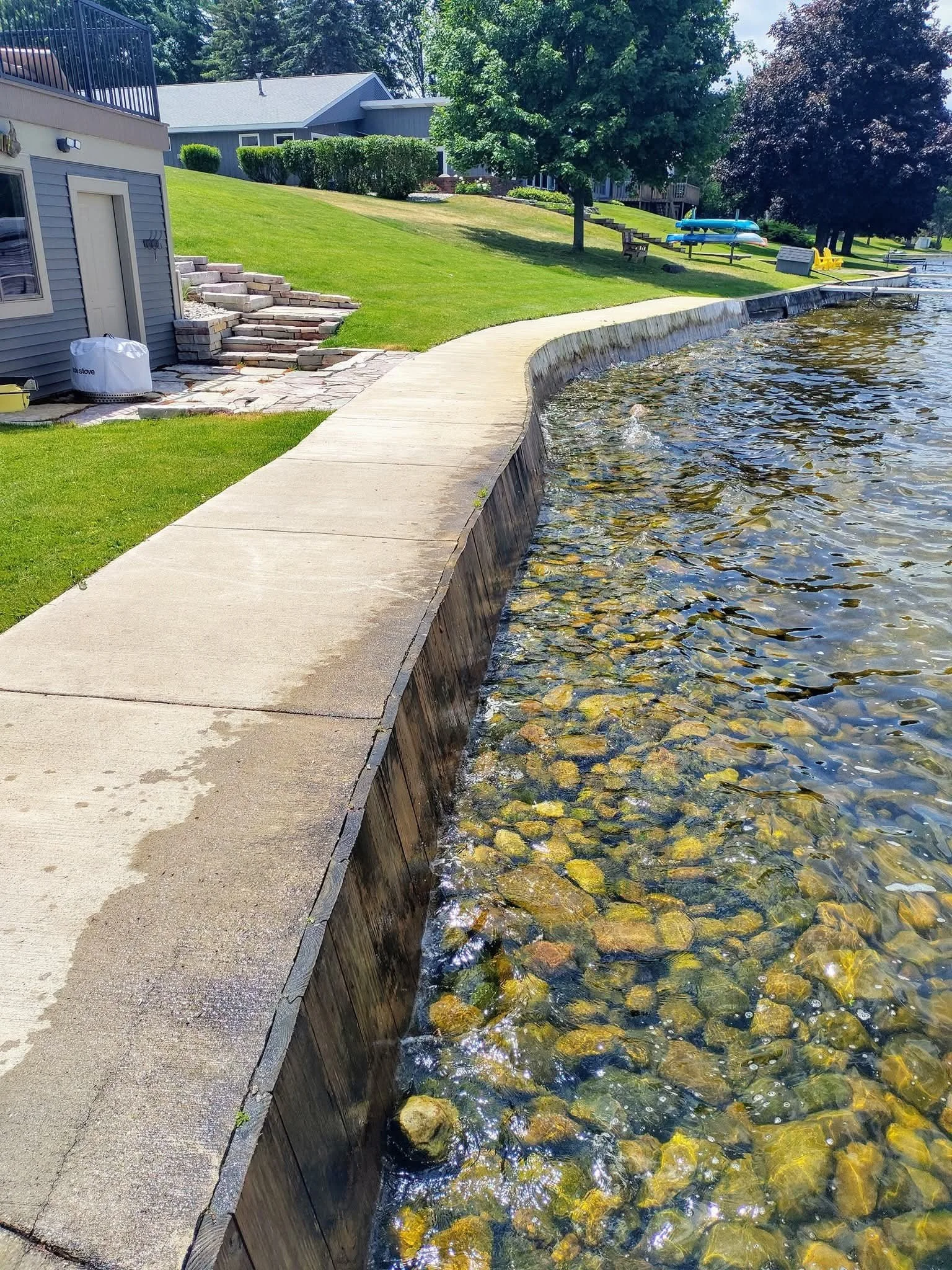 A waterfront park with a paved walkway along a rocky shoreline, lush green grass, trees, and houses in the background. There are kayaks and benches visible in the distance.