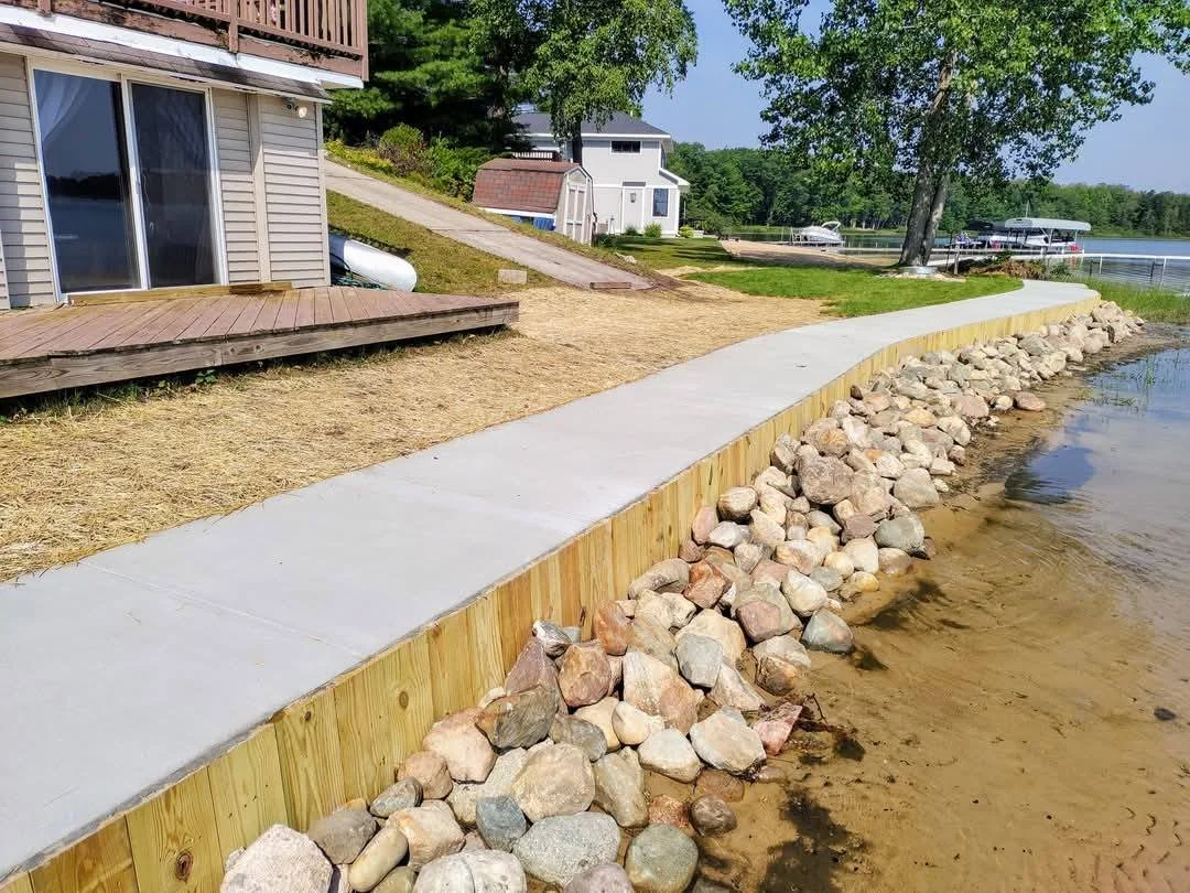 Concrete pathway along a lakeshore with rocks on one side, grass and trees in the background, and houses near the water.