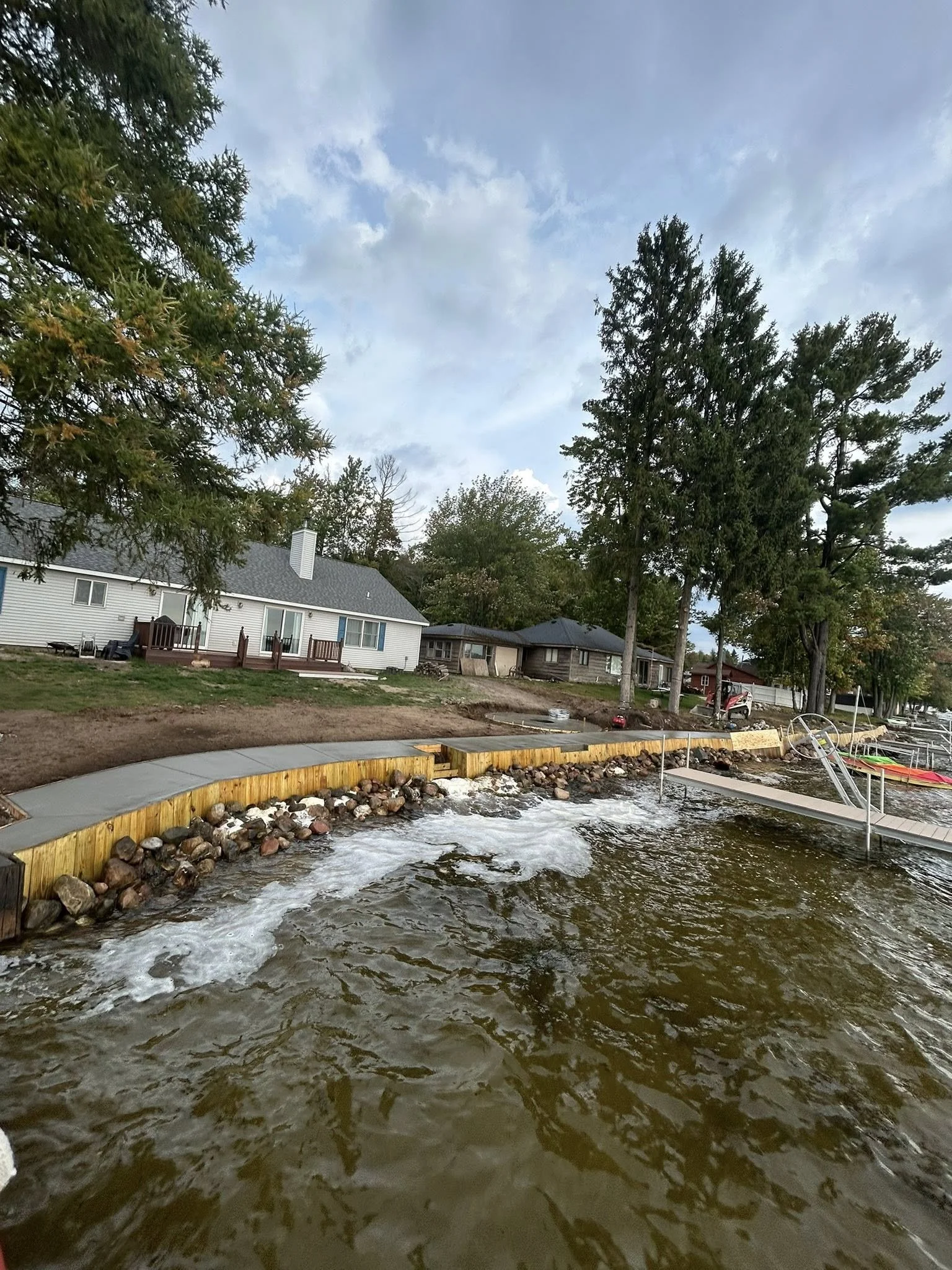 A backyard with a body of water in the foreground, wooden retaining walls, and several houses with large trees in the background. The sky is partly cloudy.