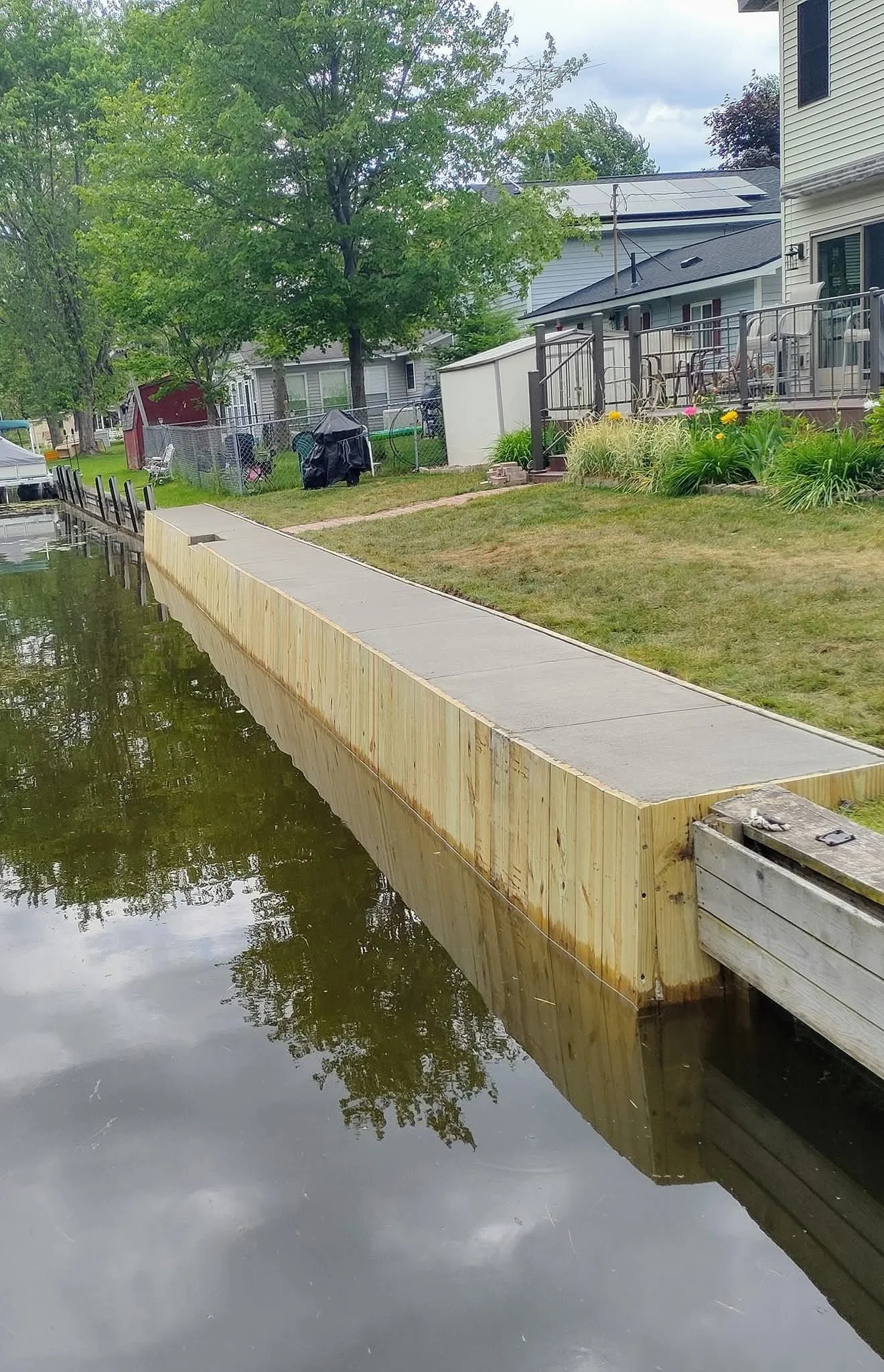 A backyard with a wooden dock extending into a pond, surrounded by grass, trees, and houses with screened porches, some with solar panels on the roofs.