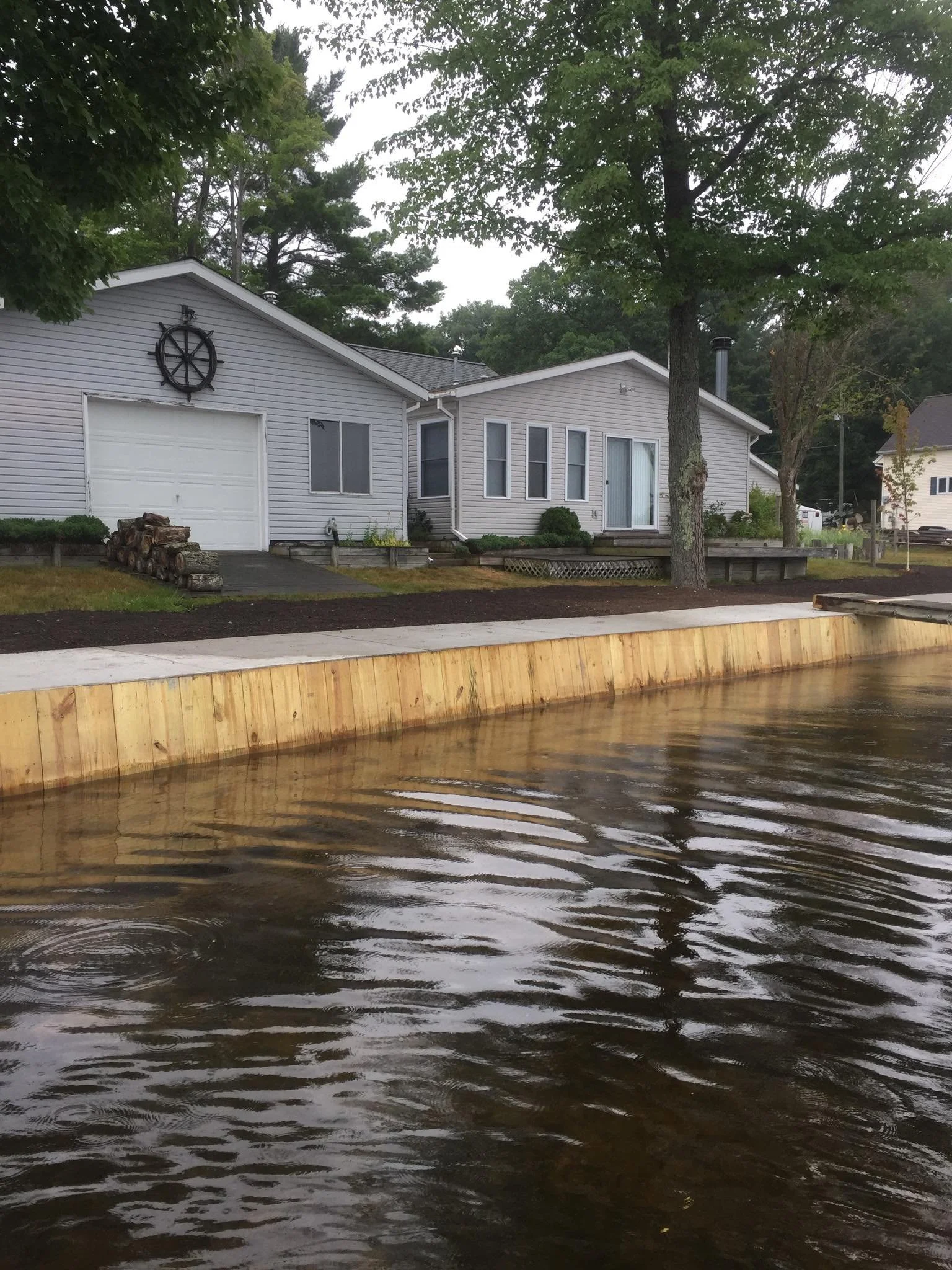 A house with gray siding and a garage, surrounded by green trees, with water in the foreground and a wooden barrier at the edge of the water.