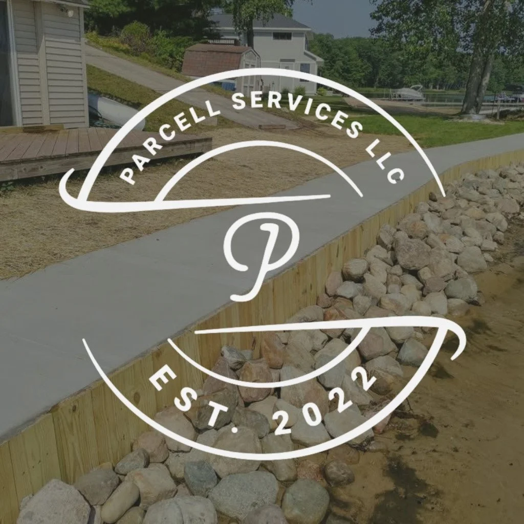 A construction site with a freshly poured concrete sidewalk or pathway, a wooden retaining wall, rocks, and a background of residential houses and a boat on the ground.