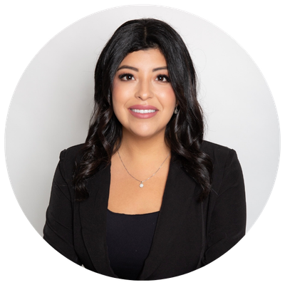 A woman with dark wavy hair, wearing a black blazer and necklace, smiling at the camera against a plain light-colored background.