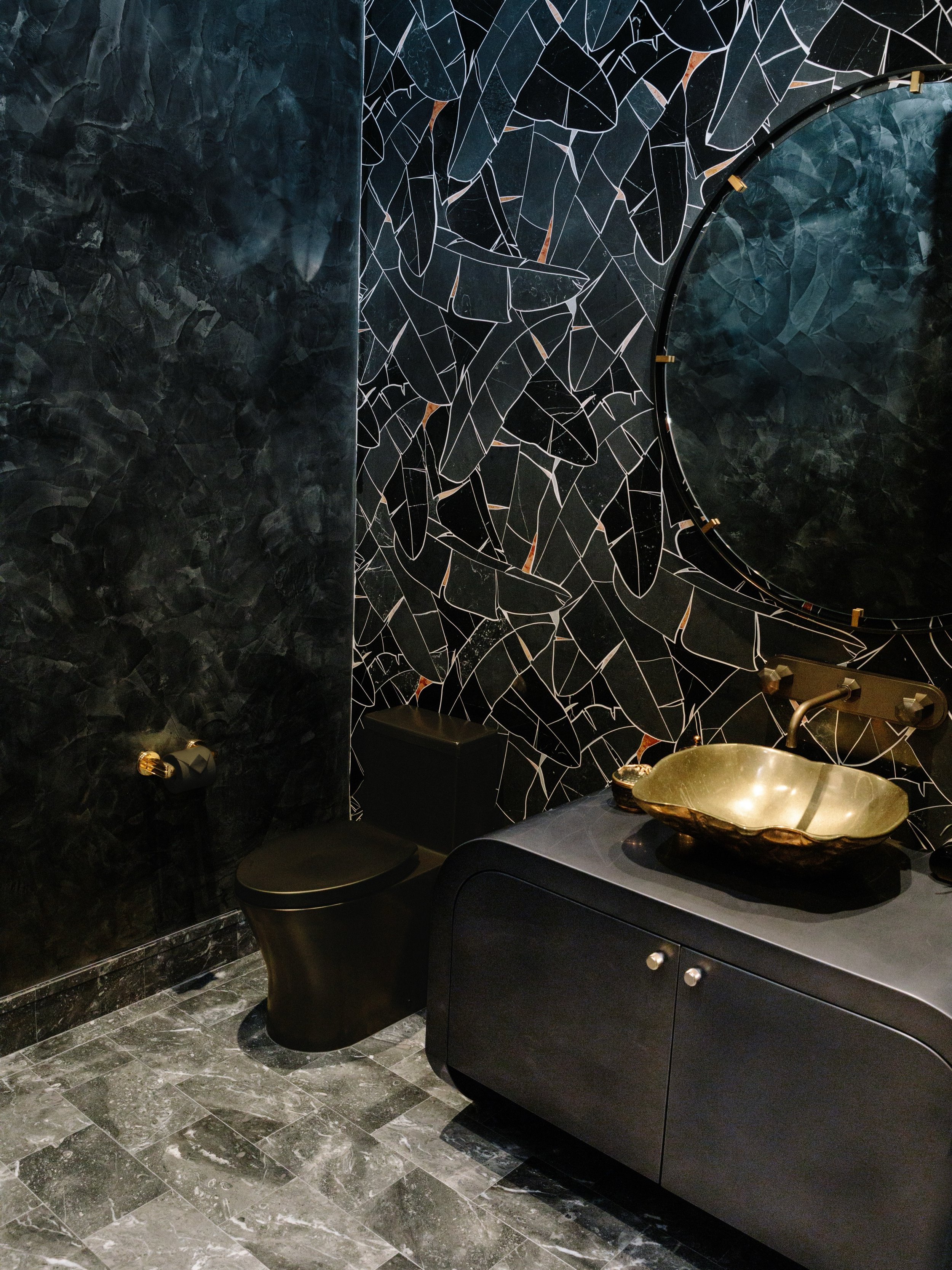 Step inside and the light changes. The walls go quiet, deep and glossy like the ocean after dark, and the marble mosaic catches you like a glance across a candlelit room. A scalloped bronze basin rests on a custom vellum-wrapped cabinet from Jimeco, 