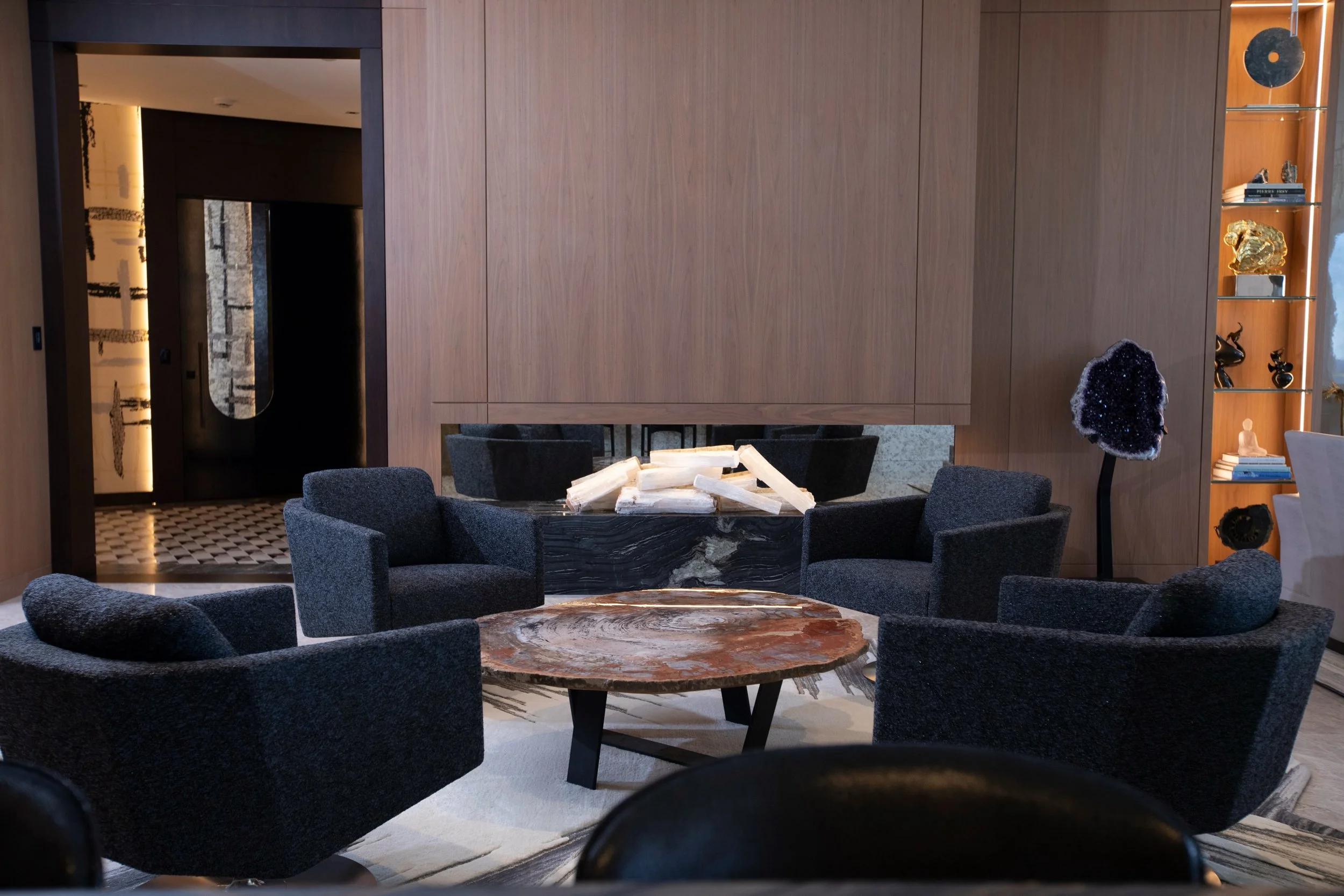 We wanted this room to feel like a place to gather at the end of a long day. Holly Hunt Jet chairs upholstered in a deep navy Elitis boucl&eacute; circle a coffee table cut from a single slab of petrified wood, its rings reading like a map of time. T