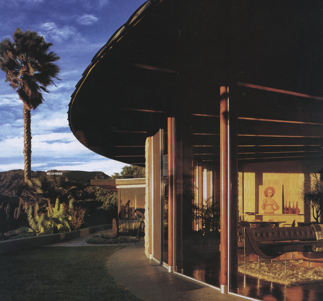 A mid-century modern house with large glass windows shows an indoor dining and lounge area with a woman statue, bottles, and plants inside. Outside, a palm tree and the Hollywood sign are visible at sunset.
