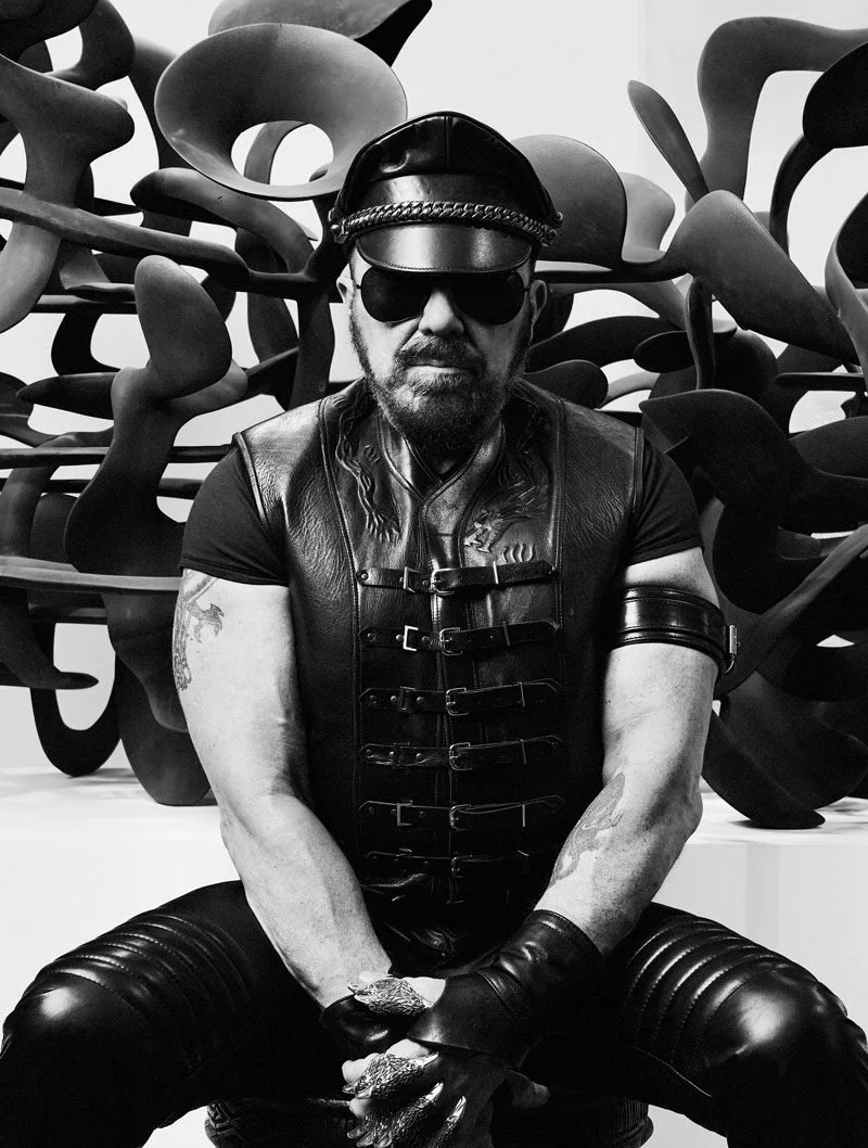 A man wearing sunglasses, a leather cap, a leather vest, and fingerless gloves sitting in front of abstract black sculptures