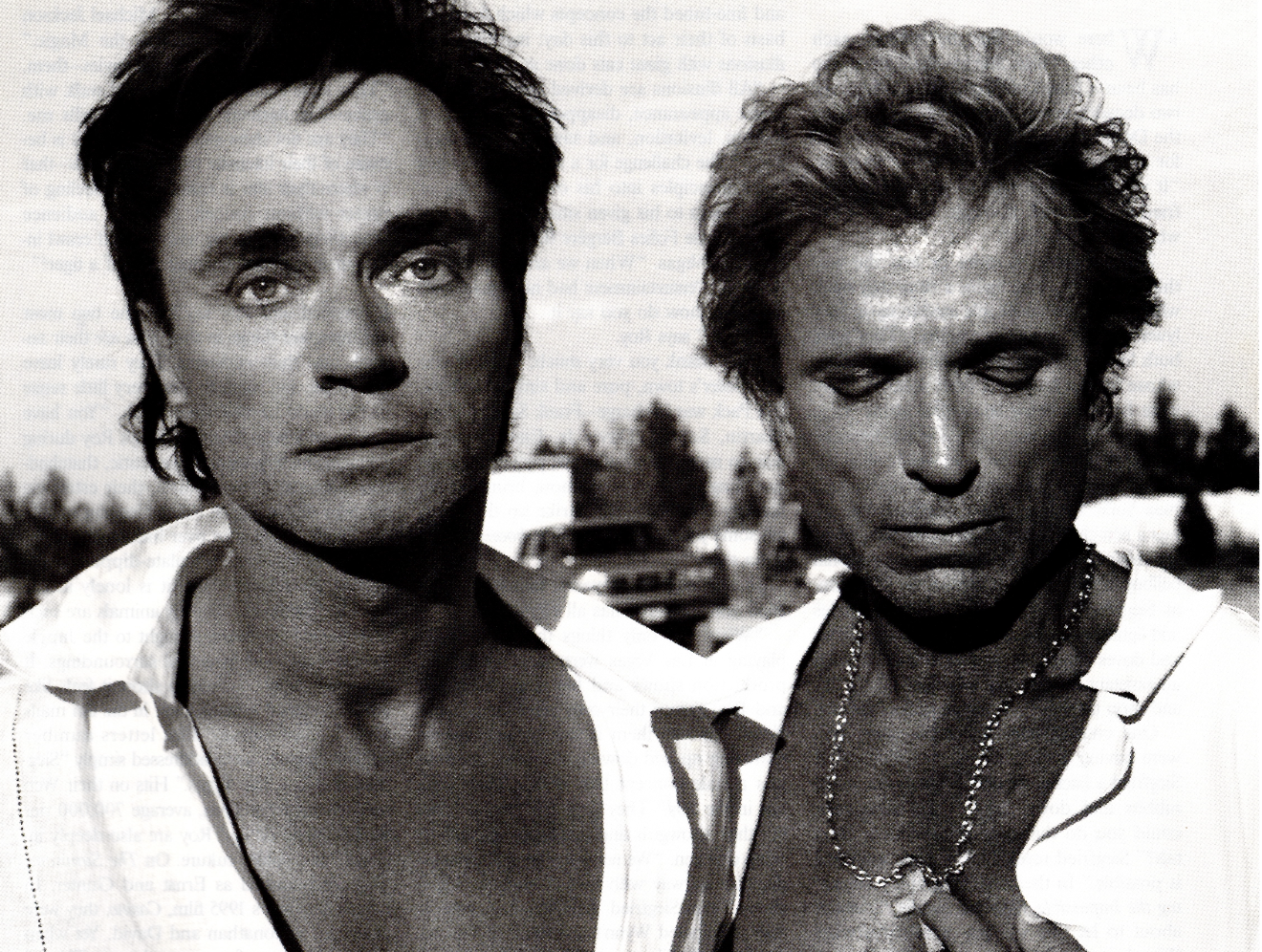 Black and white photo of two men standing outdoors, one with eyes closed and the other with eyes open, with cars and trees in the background.