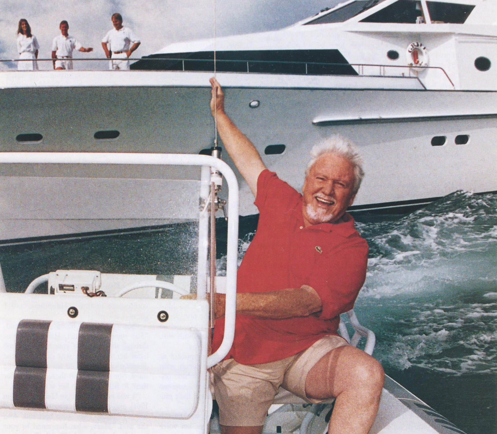 A man with white hair and a beard laughing on a boat, wearing a red polo shirt and beige shorts, with a yacht in the background and three people on the yacht.