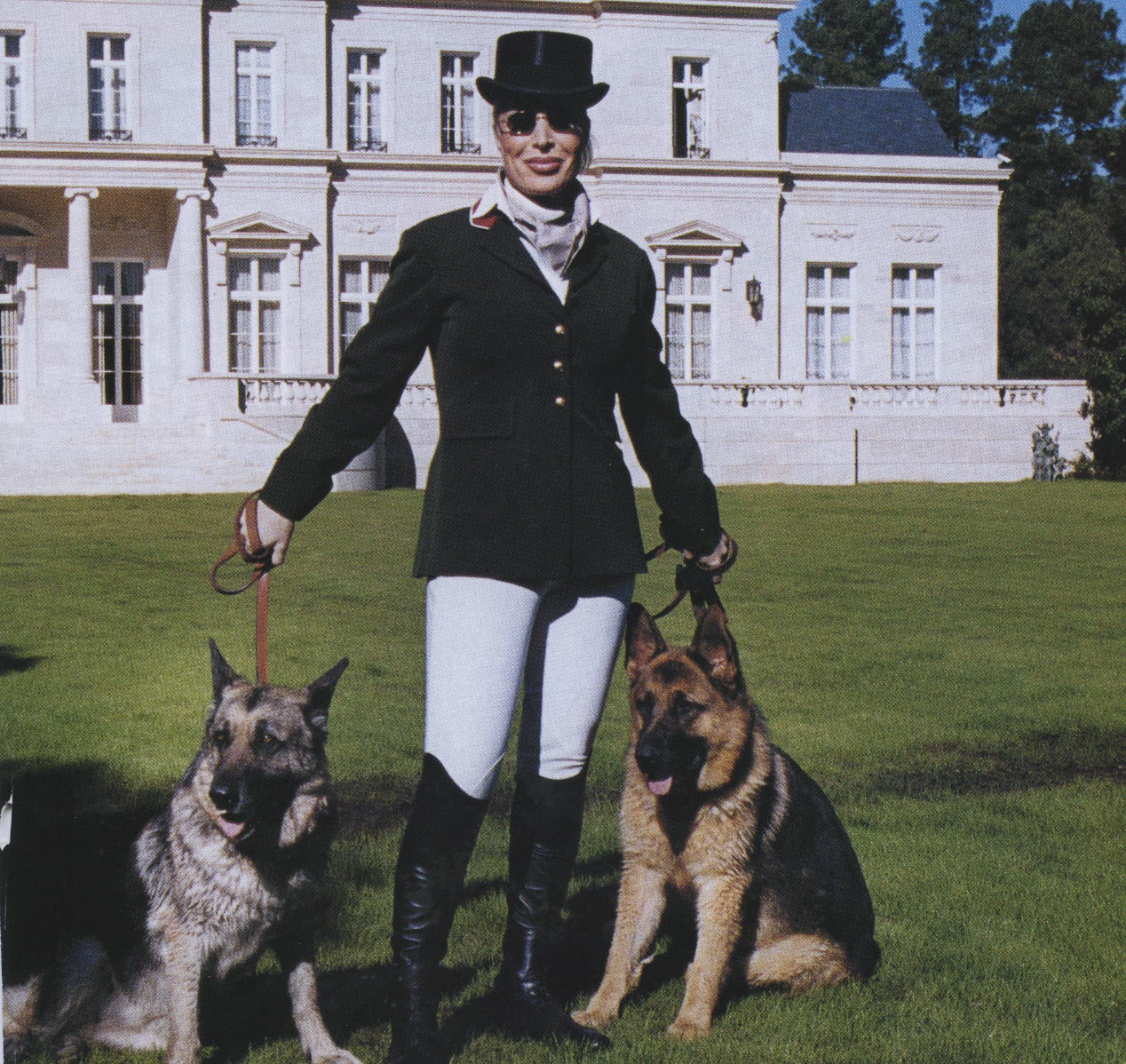 A woman in a formal riding outfit with a top hat, sunglasses, and riding boots, standing outdoors on a lawn with two German Shepherd dogs, in front of a large white mansion.