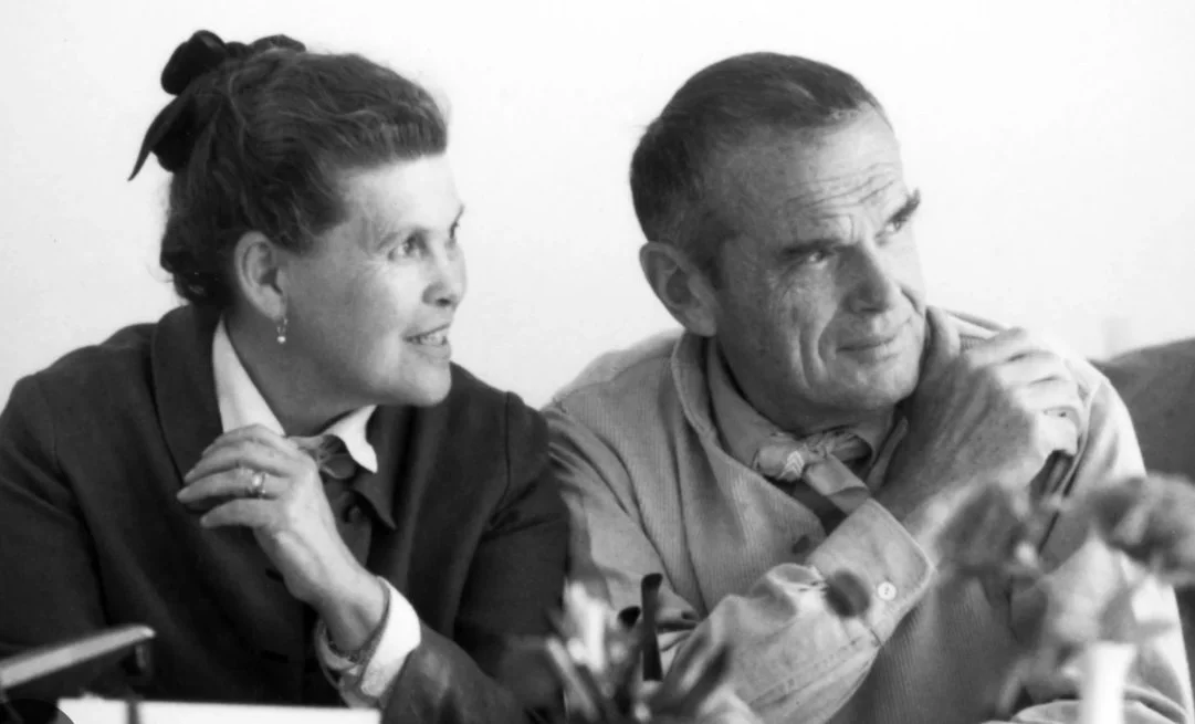 A black and white photo of a woman and a man sitting at a table, engaged in conversation. The woman is smiling and looking at the man, who appears to be listening intently.