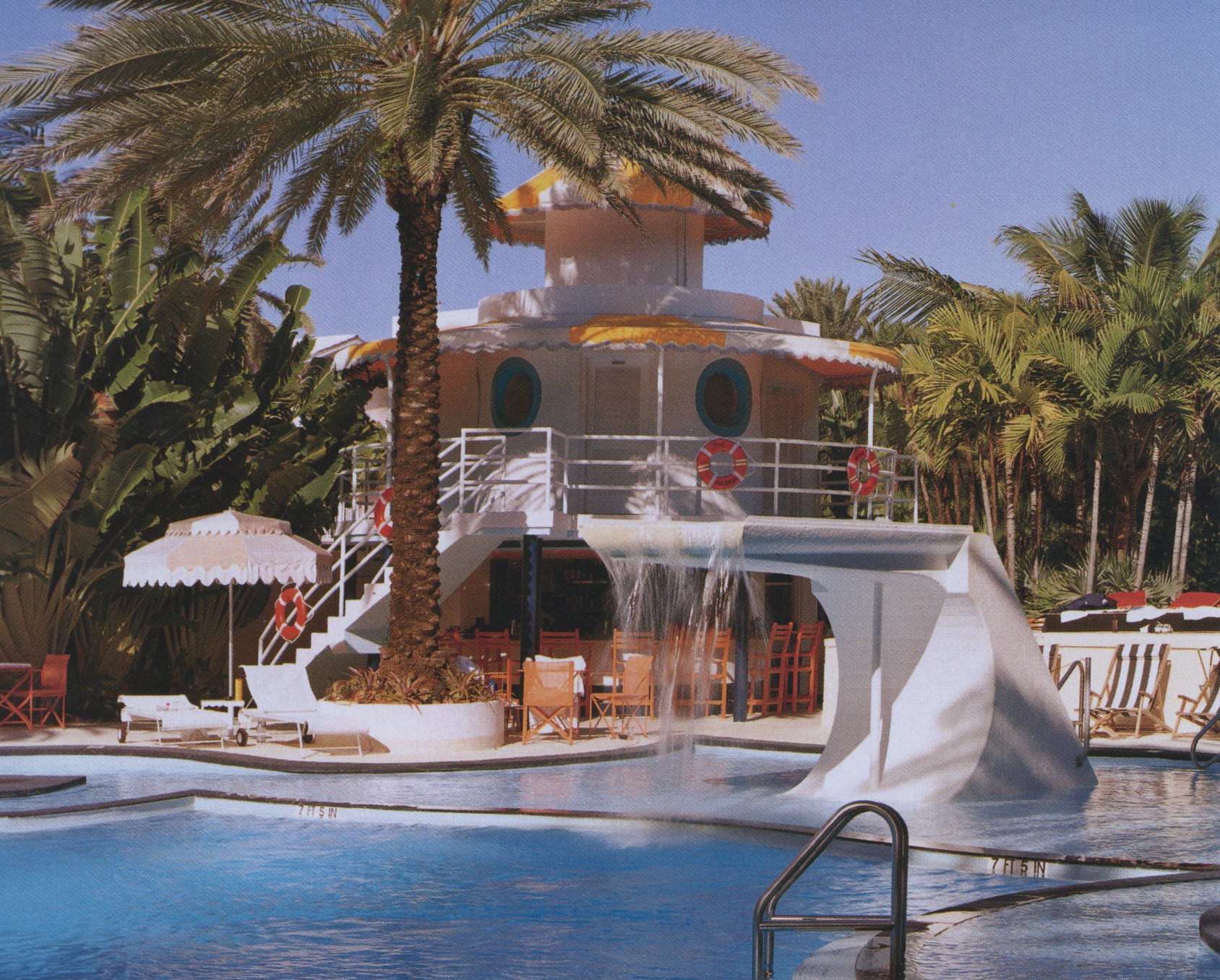 A swimming pool area with a large water slide, surrounded by lounge chairs, umbrellas, and tropical palm trees under a clear blue sky.