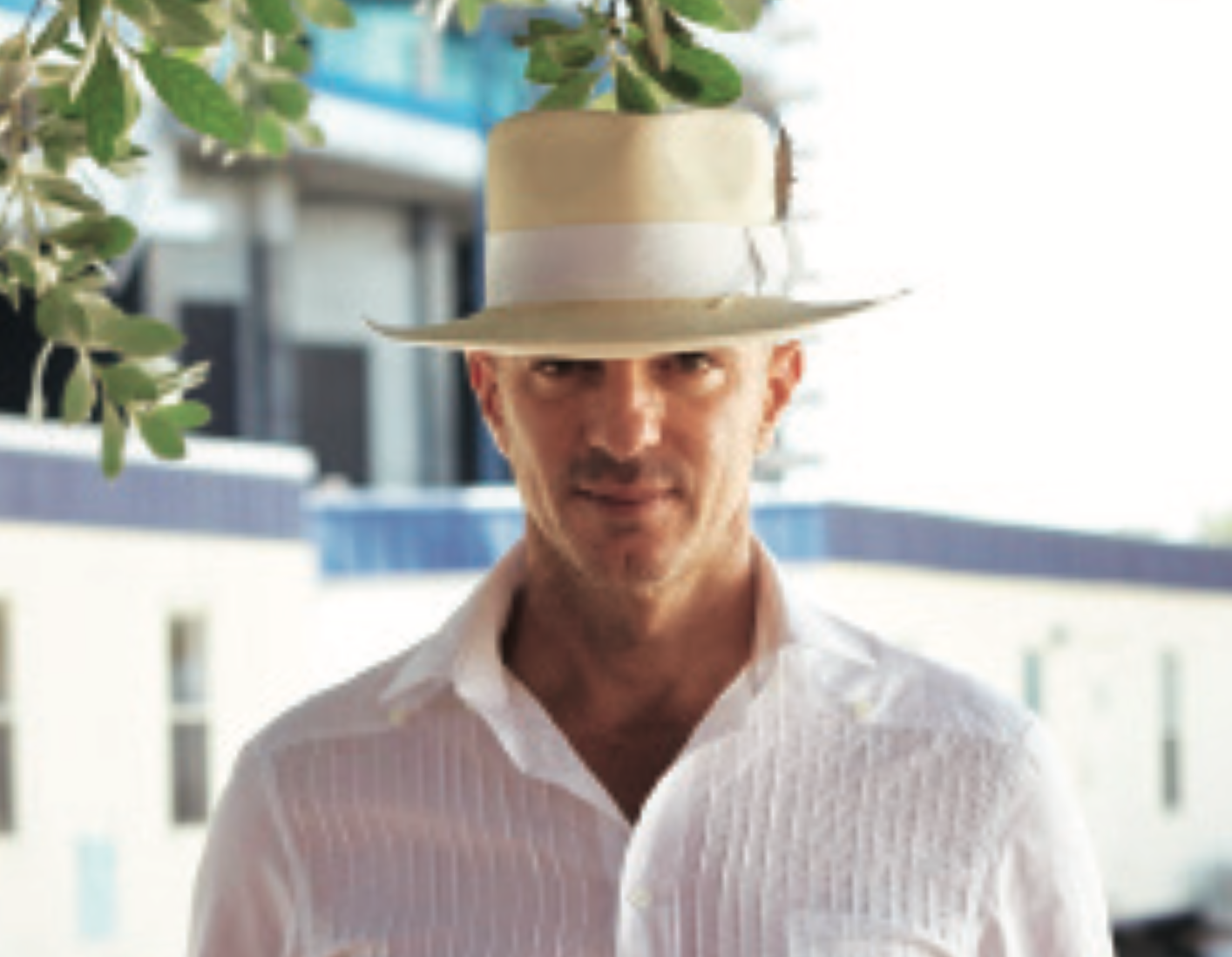 A man wearing a white shirt and a wide-brimmed straw hat with a ribbon, standing outdoors with buildings and greenery in the background.