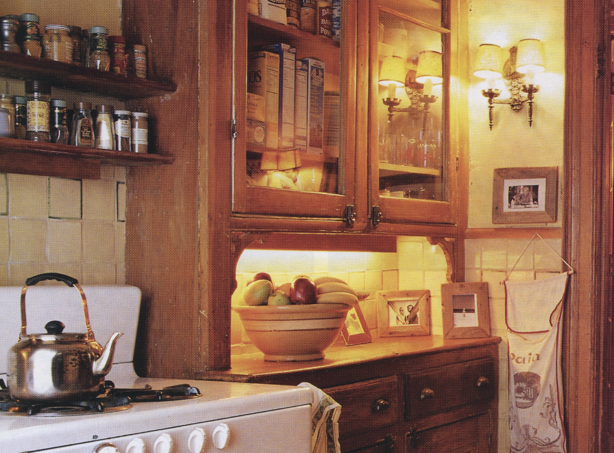 A cozy kitchen corner with a white stove, a silver kettle on the stove, and wooden cabinets with glass doors. Inside the cabinets are food boxes. On the counter, there is a bowl of apples. The area is warmly lit with yellow lighting, and there are framed photos and a hanging cloth bag on the wall. A wall sconce with two lamps is also visible.