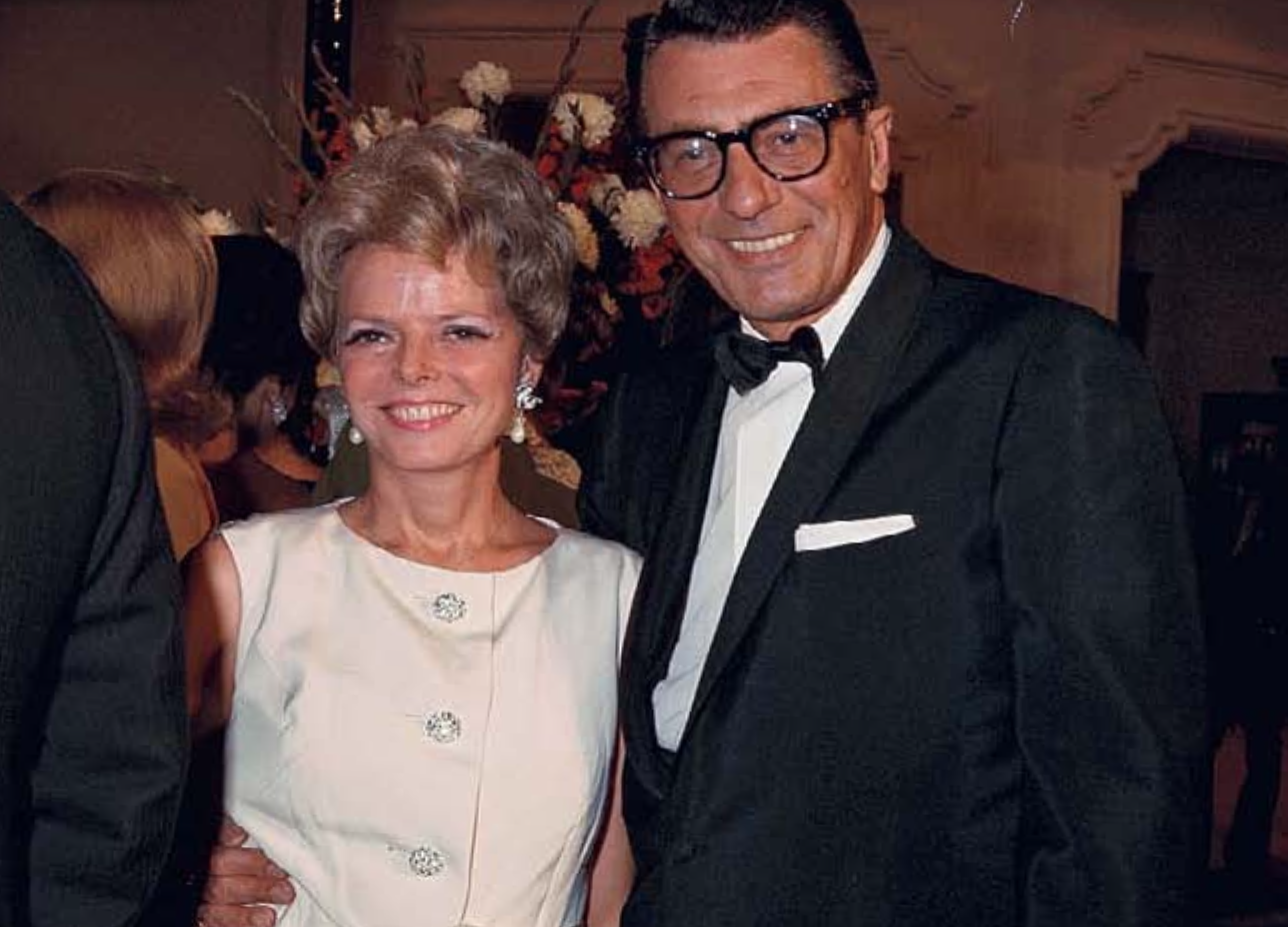 A smiling older woman in a white sleeveless dress with jewelry, standing next to a smiling man in a black tuxedo and glasses, at a formal event.