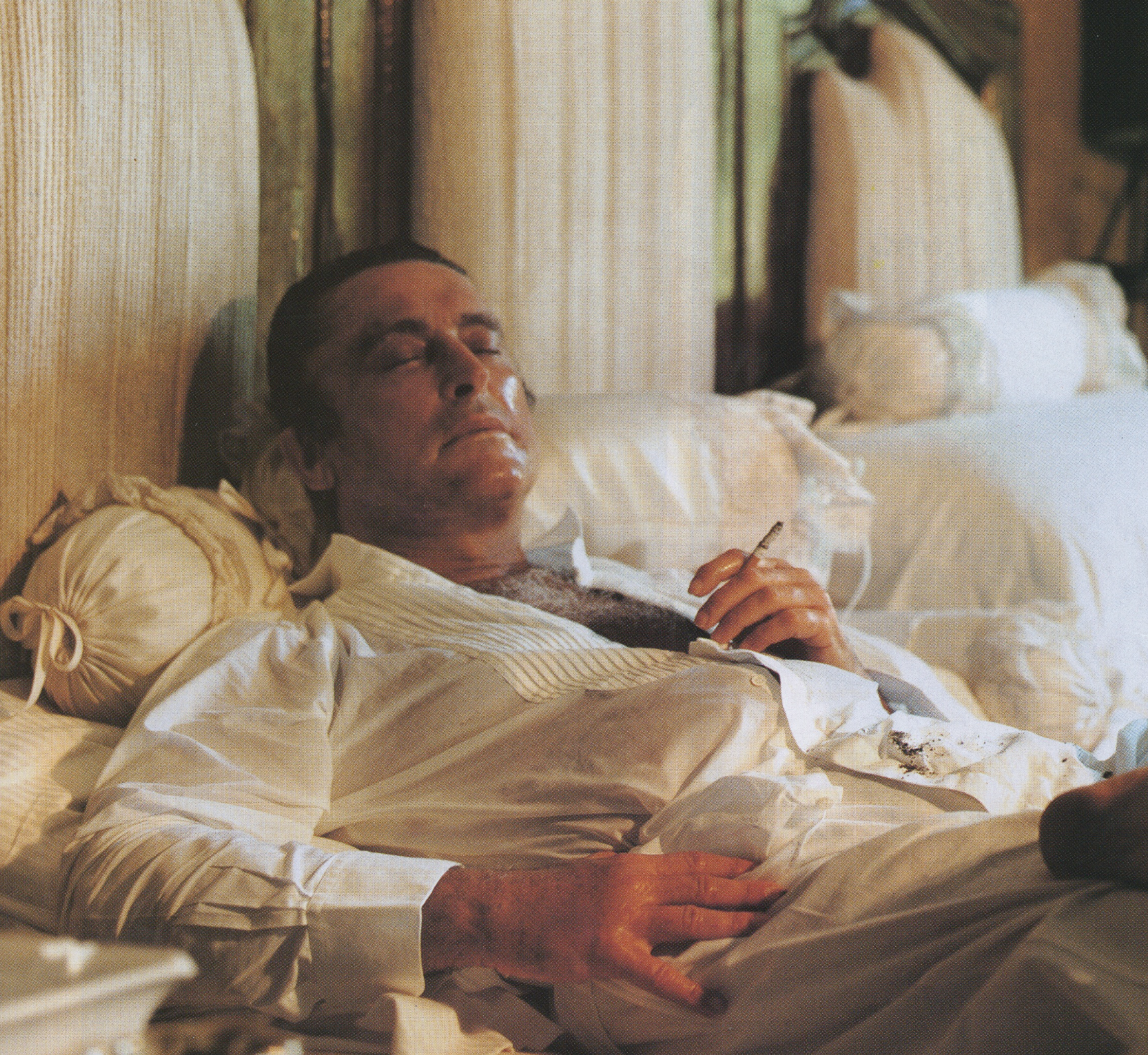 A man lying on a bed with eyes closed, dressed in a white shirt, holding a cigarette in his right hand, in a warmly lit room with cream-colored walls and upholstered headboard.