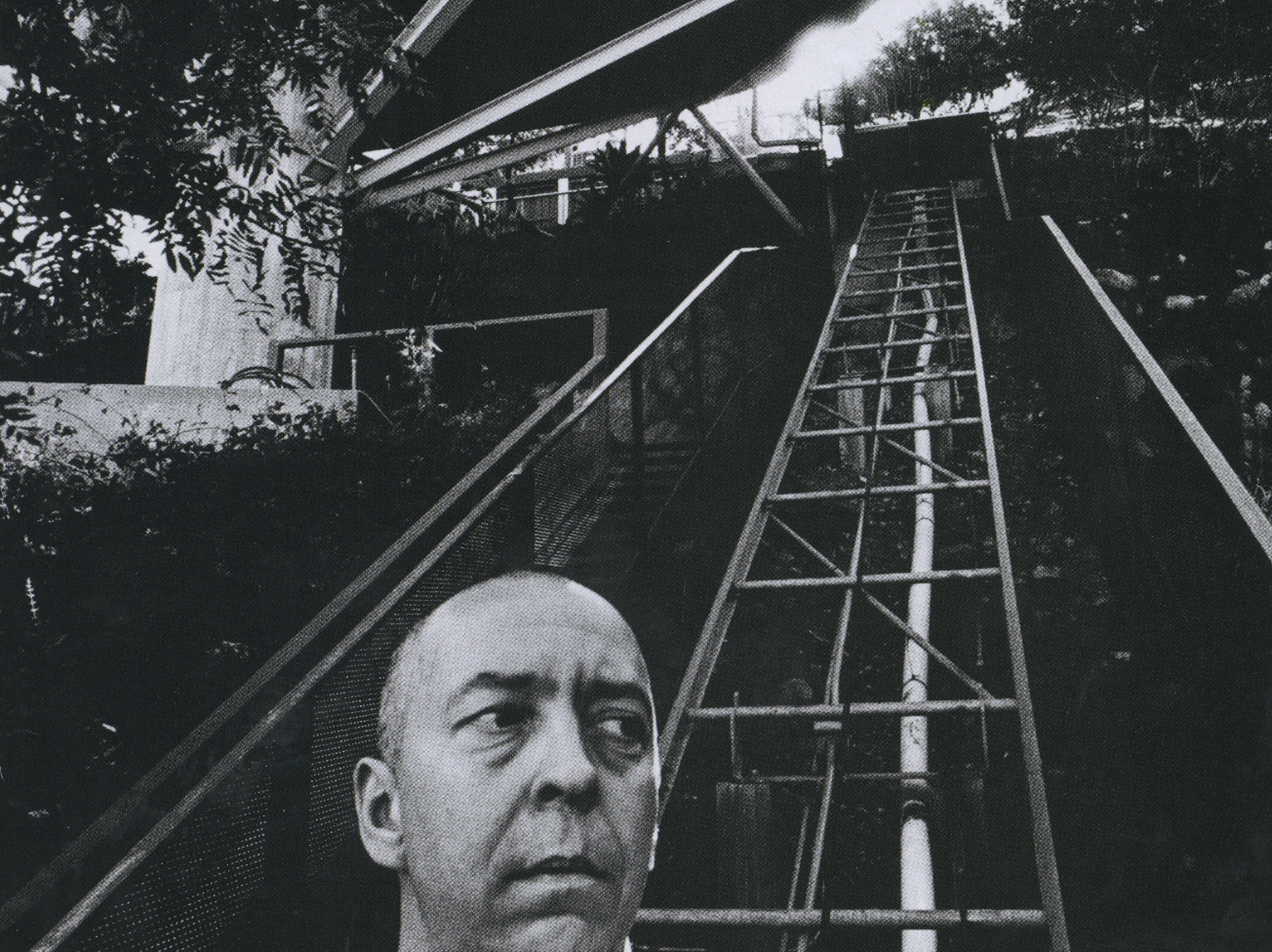 Black and white photo of a man with a serious expression standing in front of a roller coaster track with a tall climbing structure, trees, and foliage in the background.