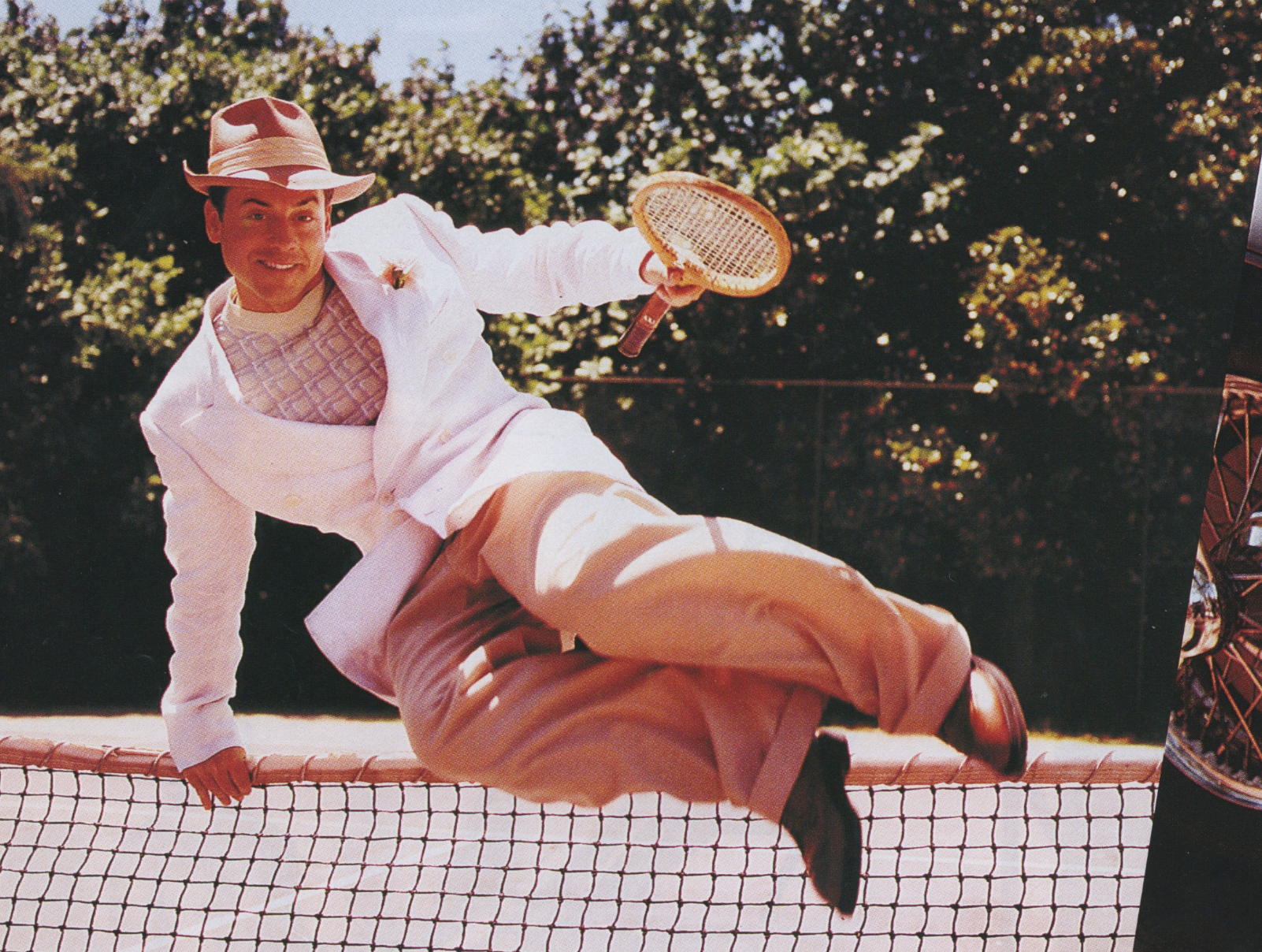 Man in a white suit and hat falling backwards over a tennis net while holding a tennis racket, outdoors with greenery in the background.