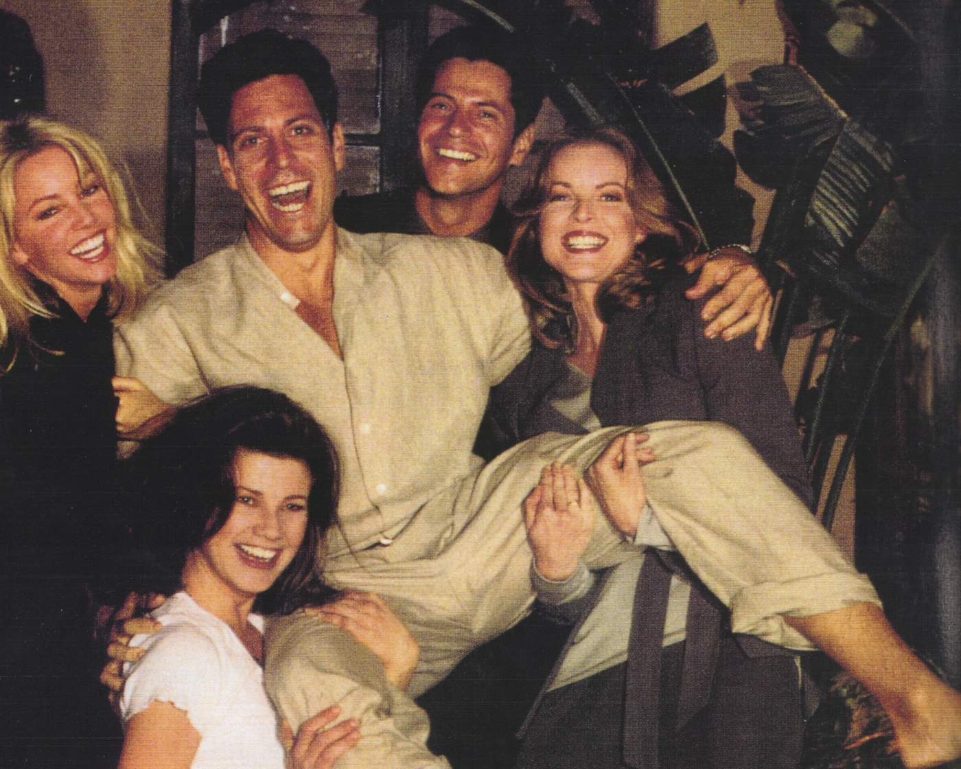 Six adults smiling and laughing while posing together for a photo. One person is being held up horizontally by the others.
