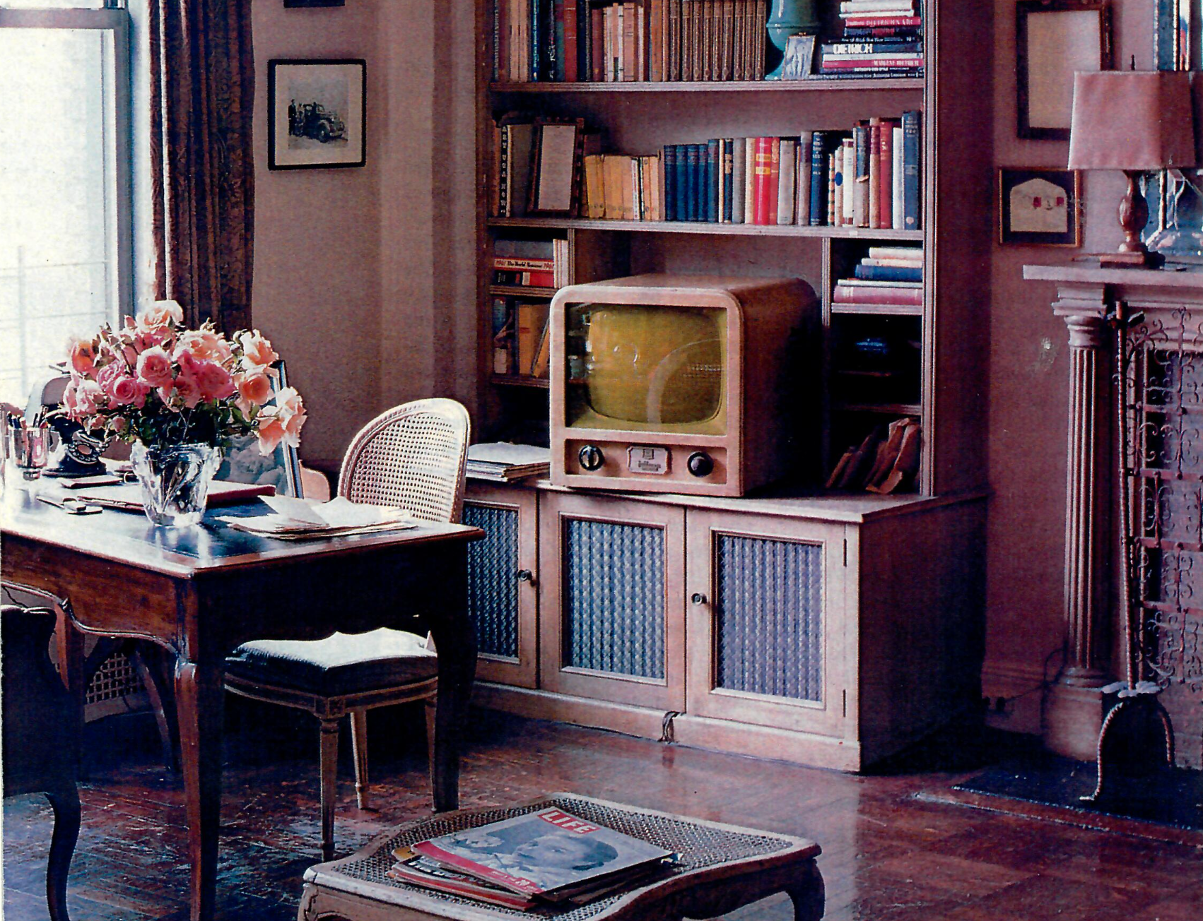 A vintage living room with a wooden bookshelf filled with books, an old television set on a wooden cabinet, a table with a vase of pink flowers, a chair, and a small bench with magazines, with framed pictures on the walls and a window with curtains.