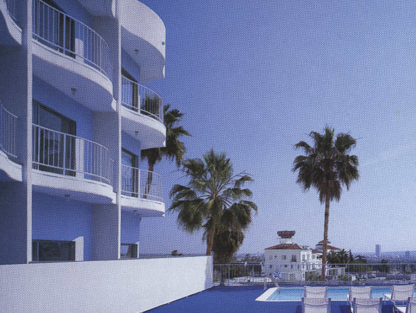 A building with white curved balconies, a swimming pool, lounge chairs, and palm trees under a clear blue sky.