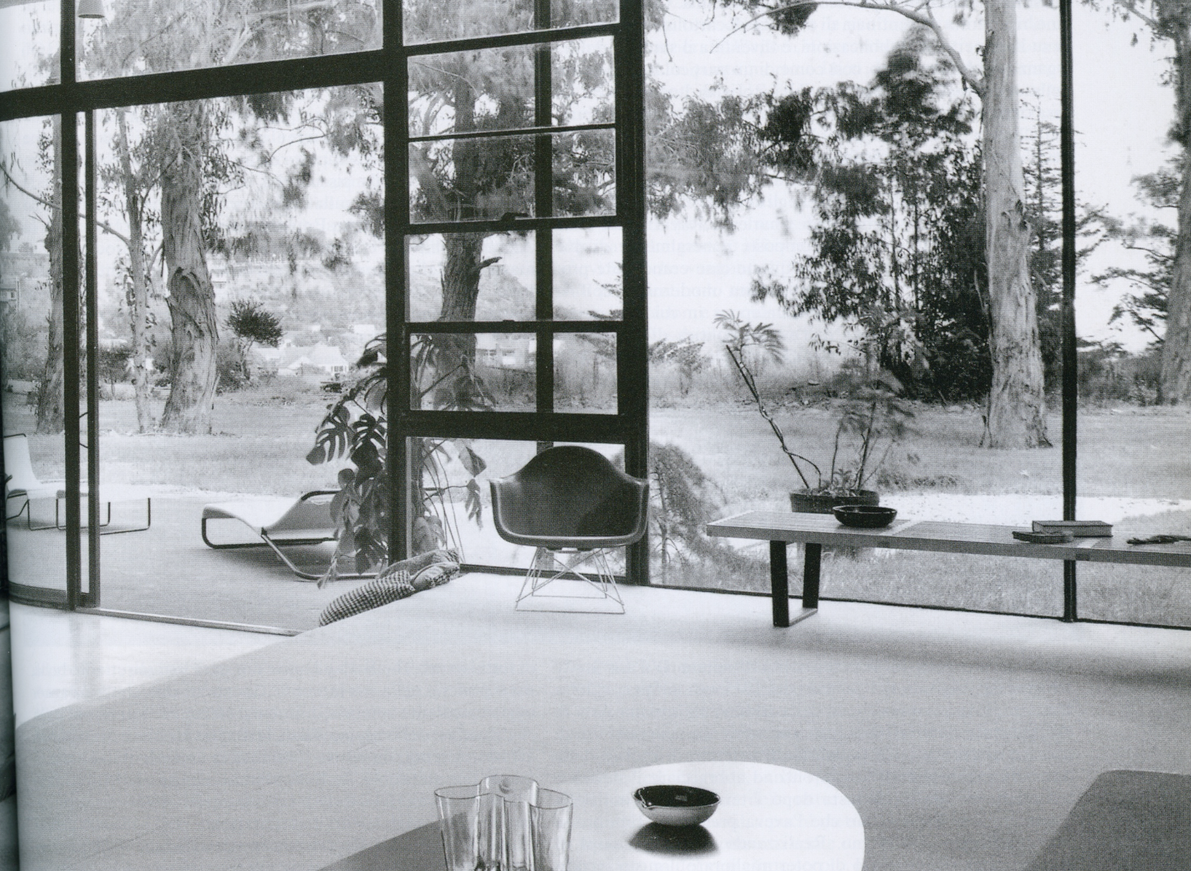Black and white photo of a room with large floor-to-ceiling windows overlooking trees and greenery outside. Inside, there is a table with a bowl and some glasses, a chair, and a bench with a bowl on it.