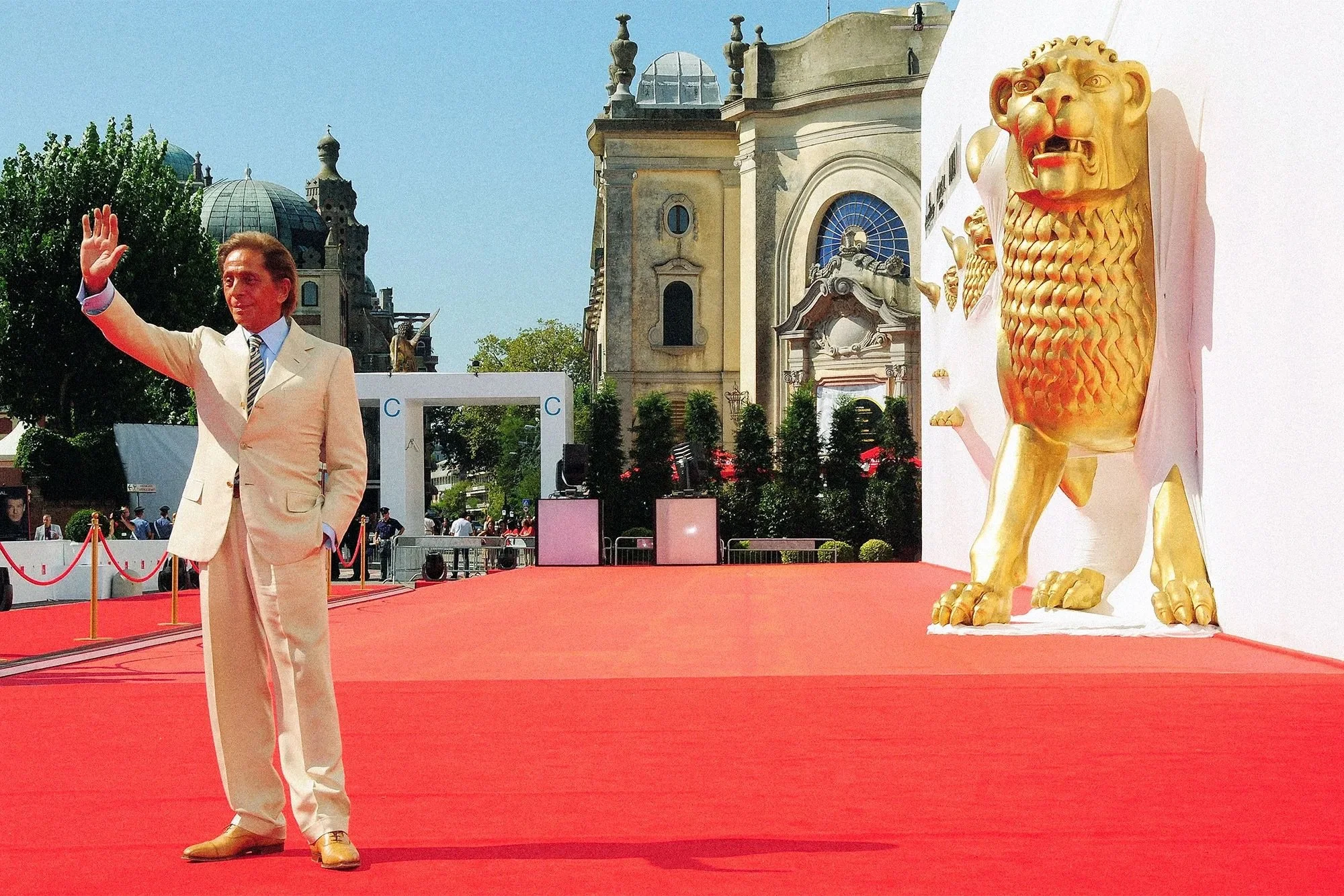 A man in a cream-colored suit and tie waving on a red carpet with a large golden lion statue to the right. The background features historic buildings and green trees under a clear blue sky.