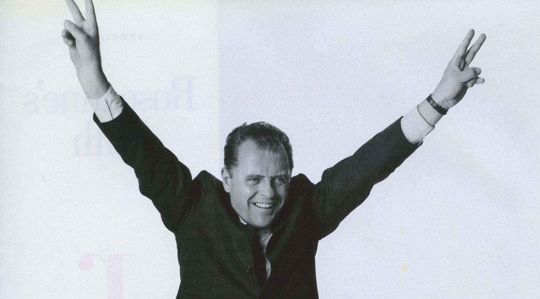 A black and white photo of a man in a suit with a tie, smiling and raising both arms with peace signs, in a celebratory pose.
