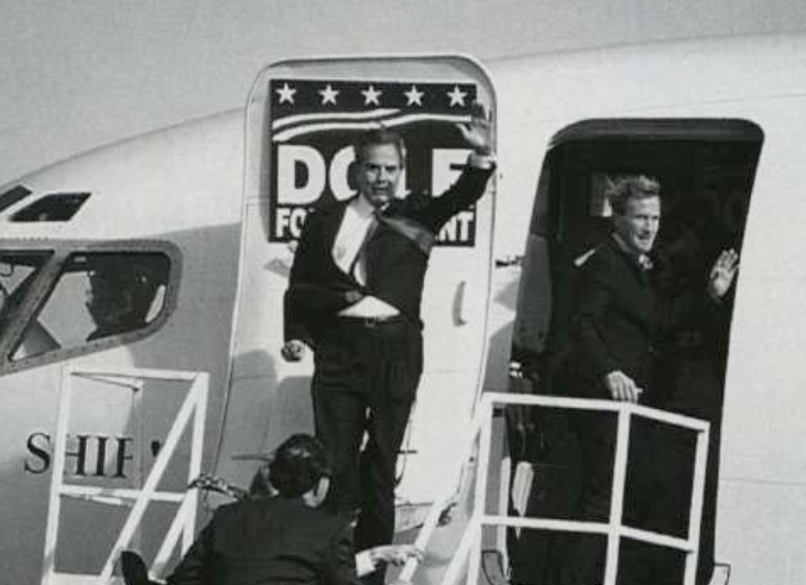 Historical black-and-white photograph of two men in suits waving from airplane stairs, with a third person below adjusting something.