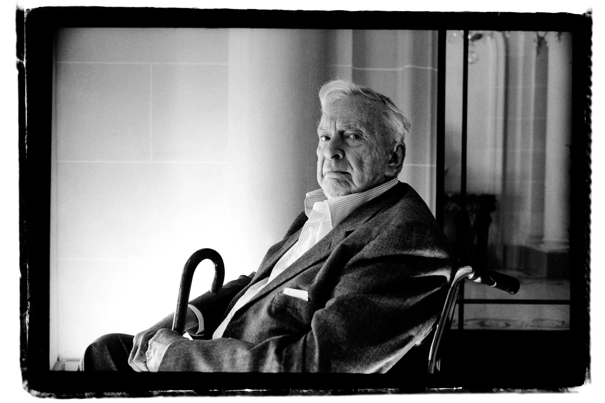 Black and white photo of an elderly man with white hair, sitting in a wheelchair, looking serious or stern. He is wearing a striped collared shirt and a jacket, with a cane resting in his hand. The background shows a tiled wall and curtains.