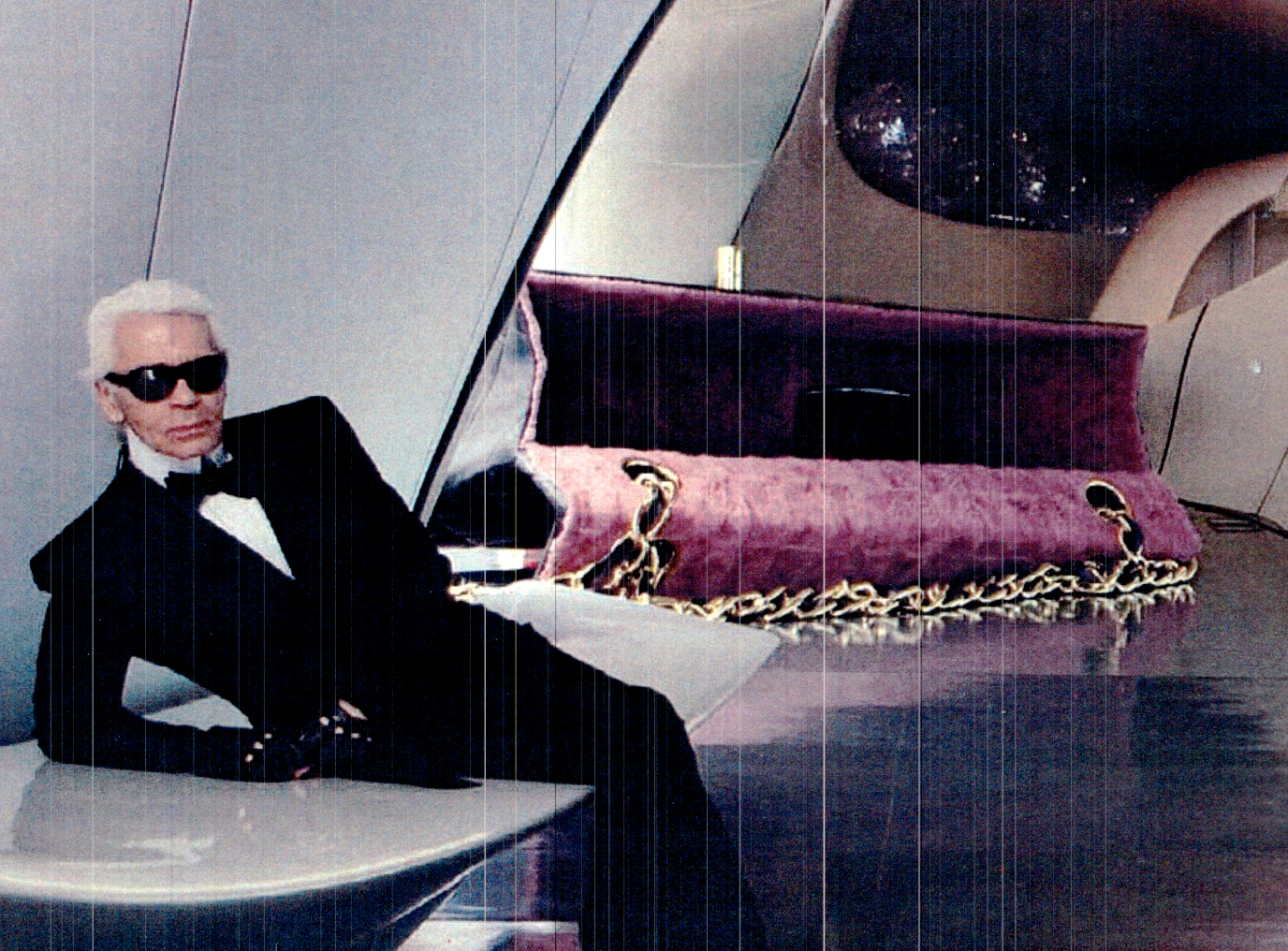 An elderly man with white hair, sunglasses, a tuxedo, and a bow tie, sitting on a bench in a modern interior space with a purple velvet couch and decorative elements.